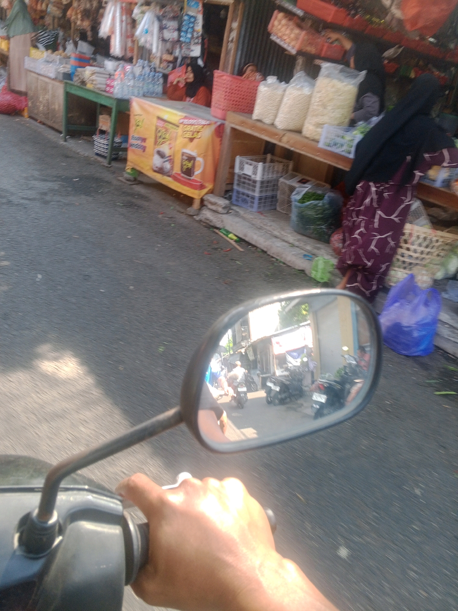 A first-person perspective shot shows a person riding a motorcycle in Wonogiri, Indonesia. The foreground features the right hand of the rider gripping the motorcycle's handlebar and throttle. The motorcycle's side mirror reflects a street scene with several parked motorcycles and some people. In the background, there is a roadside market with various stalls selling goods, including food items and household products. The market stalls are made of wood and covered with tarps. Bags of produce and packaged goods are visible. The scene appears to be during daylight hours.