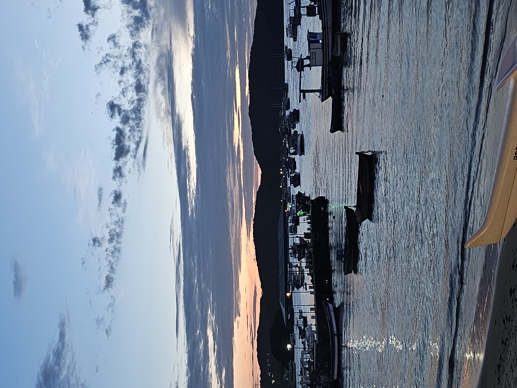 Captured at twilight, the scene unfolds in Armação dos Búzios, Brazil, showcasing a harbor filled with fishing boats moored to docks. The dark silhouettes of mountainous terrain act as a backdrop under a sky transitioning from blue to orange. Clouds are spread across the sky. The water reflects twinkling lights and the day's fading sunlight. Part of the foreground shows the side of a boat, indicating the vantage point.