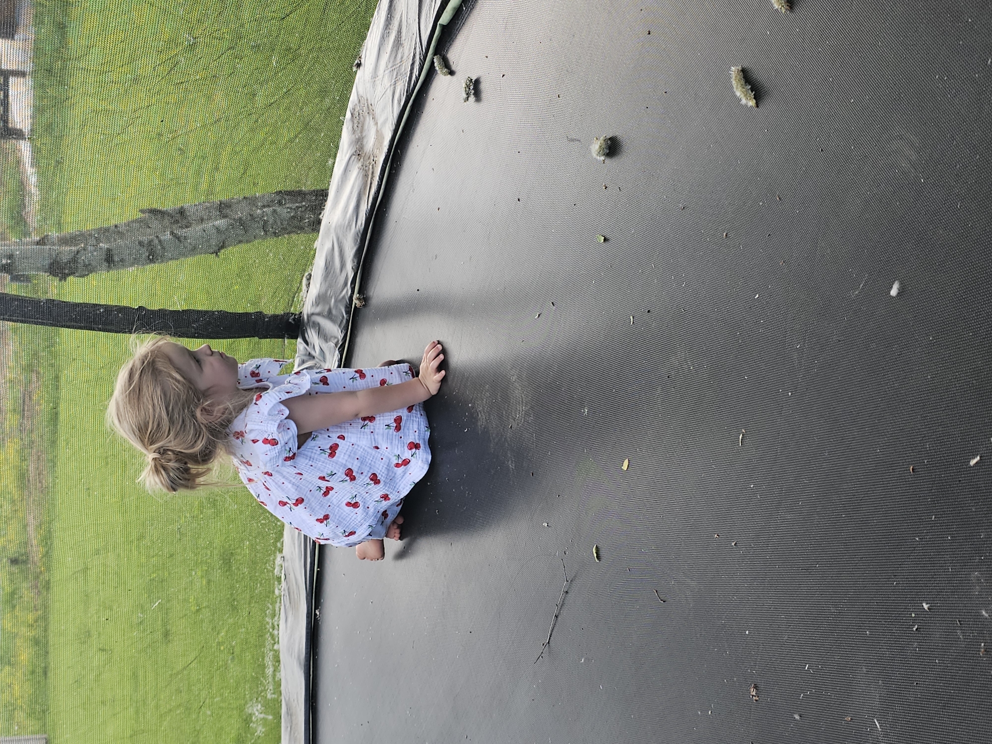 A young child with blonde hair tied in a low ponytail sits on a black trampoline surrounded by a safety net. The child wears a white dress patterned with red cherries. Outside the trampoline net, green grass is visible. There are multiple caterpillars scattered across the surface of the trampoline. The location, Мышкино, Russia, is not discernible from the image itself, but is included as part of the prompt.