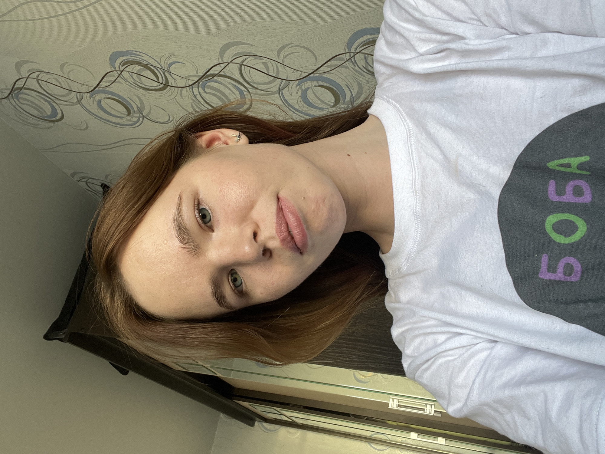 A portrait of a young woman with fair skin and auburn hair, slightly turned toward the camera. She has light green eyes and a neutral expression. She is wearing a white long-sleeved shirt with the word "BOBA" printed on it, the "B" in green and "A" in purple. The background is a wall with decorative swirls and a portion of a window frame and a dark-colored shelf or piece of furniture on the lower left side. The picture was likely taken in Chaikovsky, Russia.