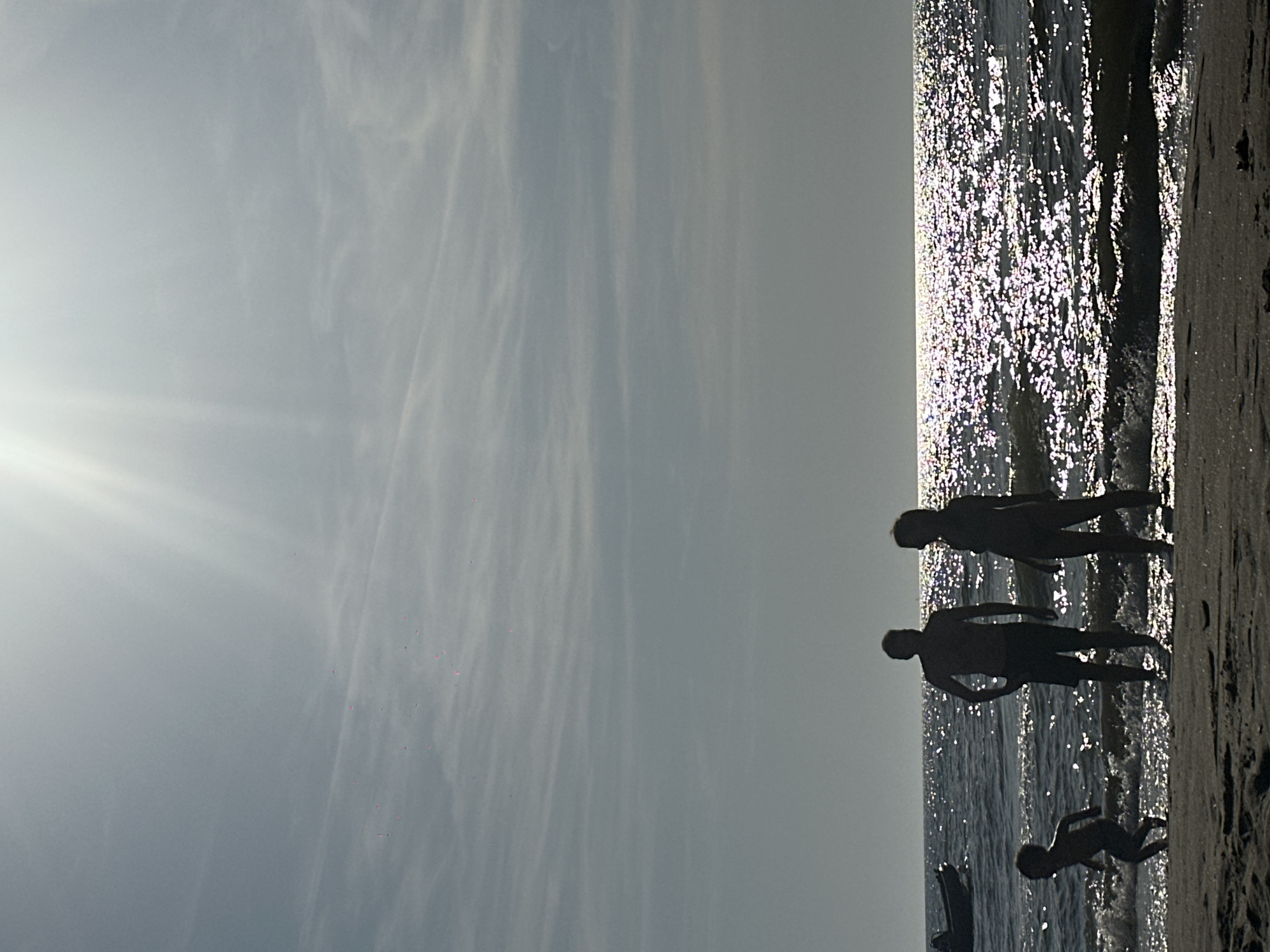 The image depicts a family silhouetted against the sun on a beach in Zandvoort, Netherlands. The sun is high in the sky, casting bright rays and creating a shimmering effect on the water. In the foreground, a family of four, two adults and two children, are standing near the water's edge. The father, wearing shorts, stands with his hands on his hips. Beside him, a woman, likely the mother, stands with her hands clasped in front of her. A young child is running in the shallow water to the left of the adults, and another child is visible in the distance on the left, near a small boat. The beach is composed of dark sand, and small waves are breaking on the shore. The ocean stretches out to the horizon, meeting a sky filled with wispy clouds.