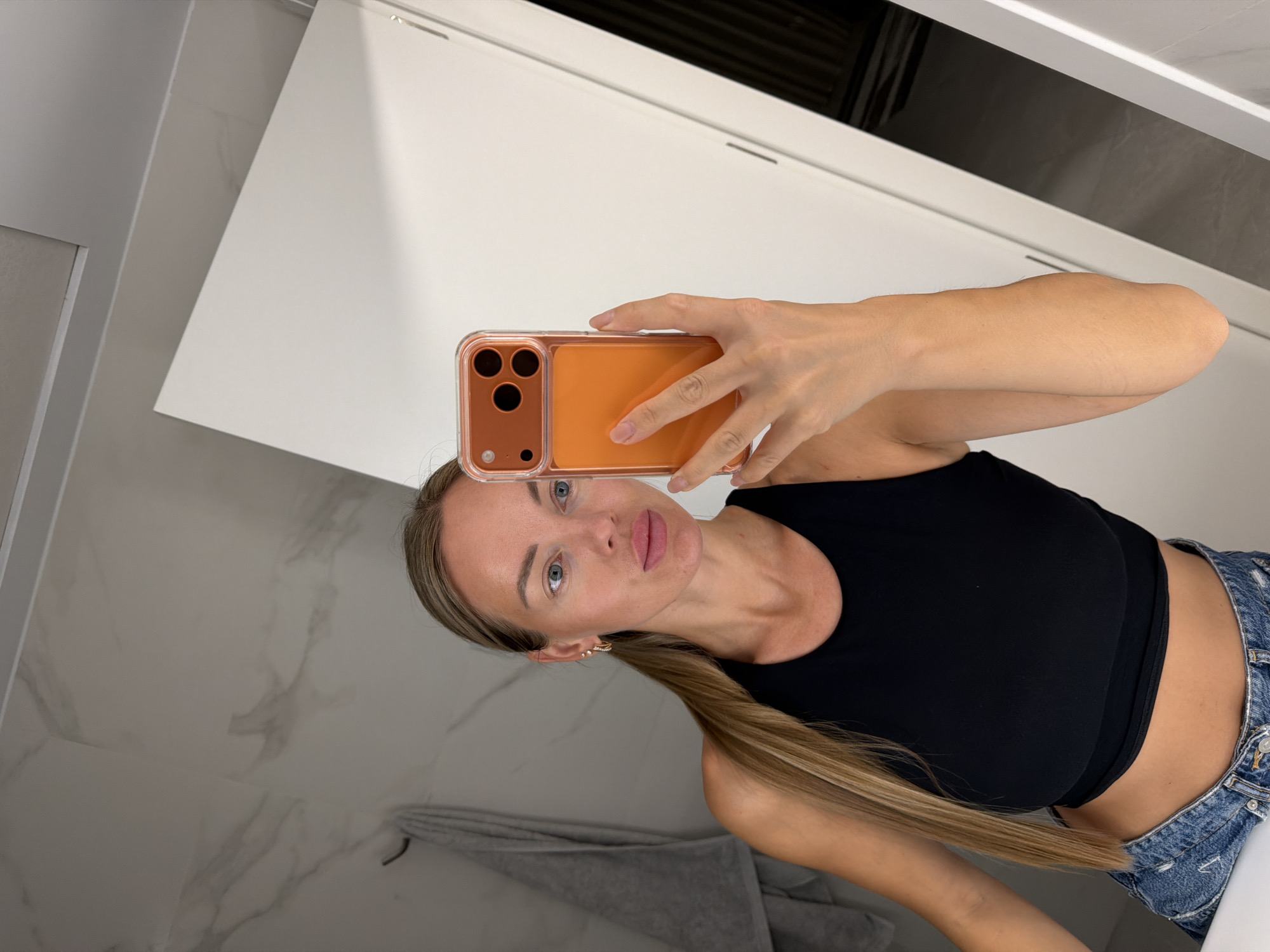 A Caucasian woman with blonde hair tied in a ponytail takes a selfie in a well-lit bathroom. She is wearing a black crop top and denim shorts. She holds up an orange-cased smartphone to capture the image. The bathroom features marble-patterned tiles on the walls and a large white cabinet to the right. A grey towel is visible hanging on a hook on the left wall. The scene is consistent with a residential bathroom, possibly in a modern apartment or hotel in Finestrat, Spain.
