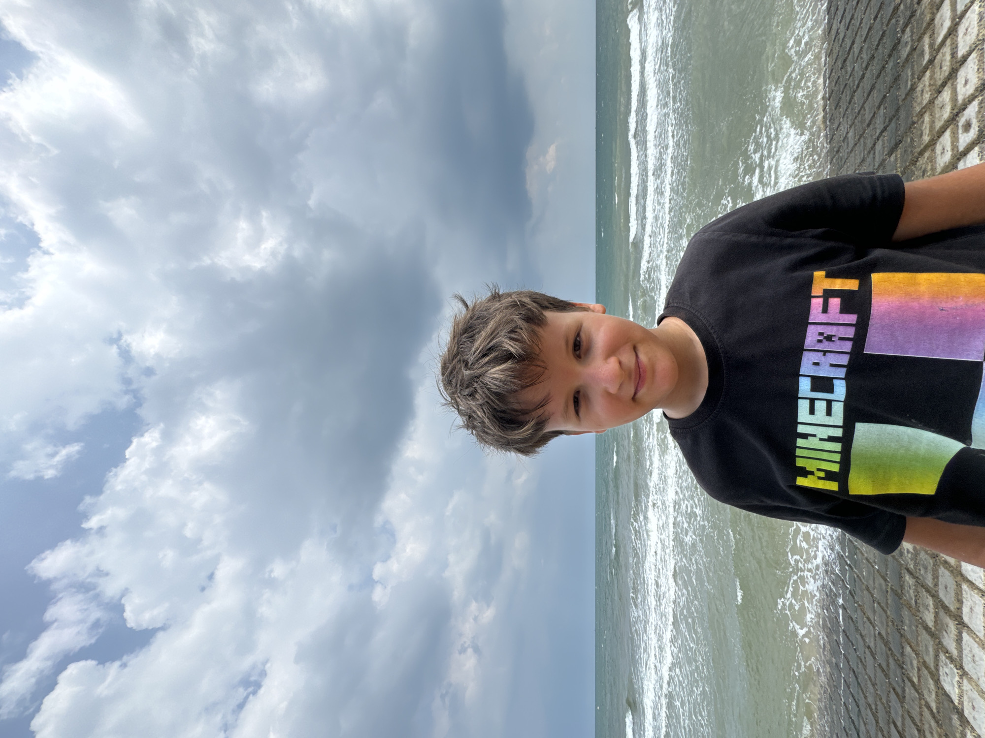 A boy wearing a black t-shirt with a colorful "Minecraft" logo stands on a tiled walkway at the edge of a body of water. The water appears to be the ocean, with waves crashing on a shore that is not clearly visible. The sky is filled with dramatic, dark clouds, suggesting an overcast or stormy day. The boy is looking directly at the camera with a slight smile. The background consists of the ocean and the cloudy sky. The location is identified as Phu Thuy Ward, Vietnam.