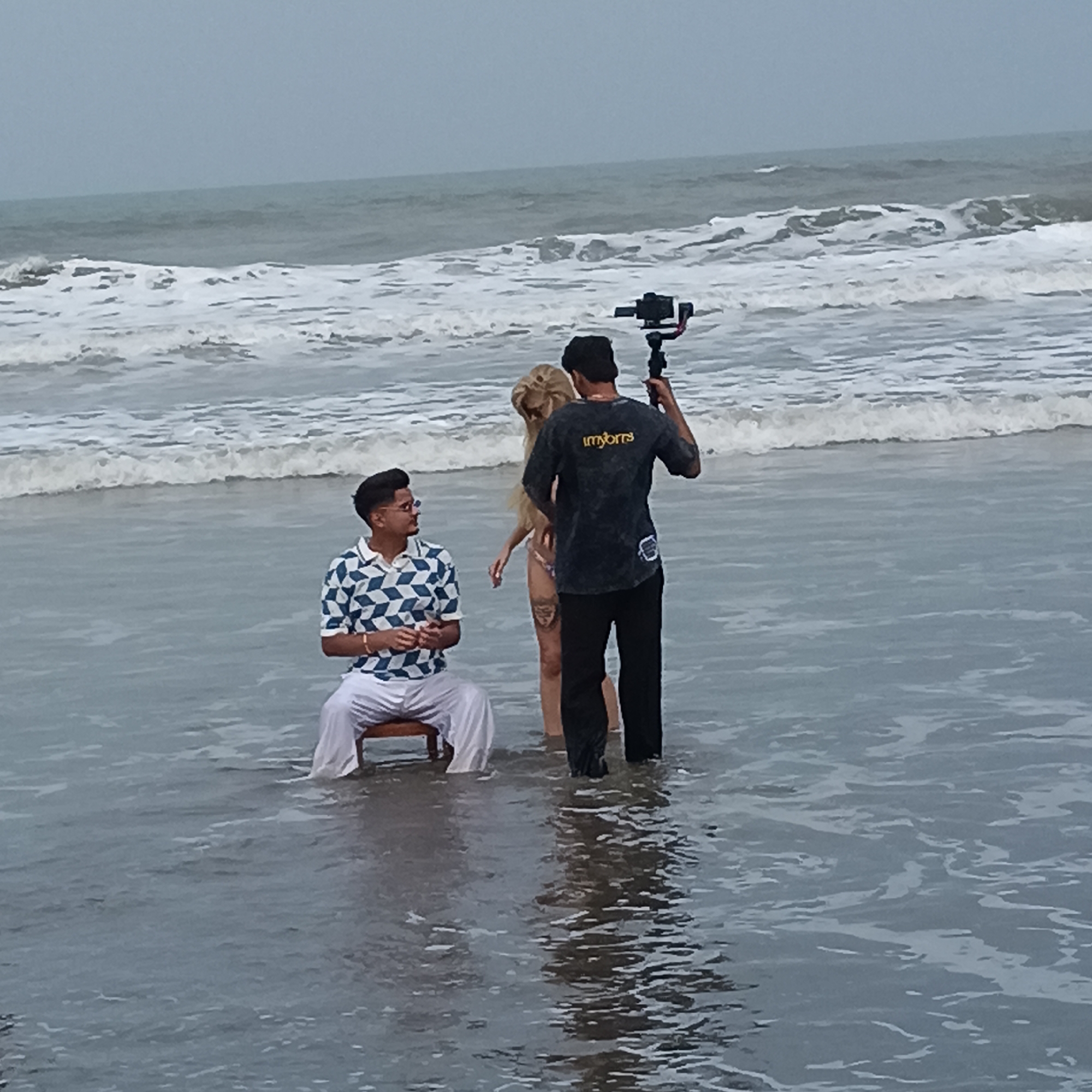 The photograph captures a scene at Mandrem Beach, India. A man sits on a wooden chair in the water, wearing a geometrically patterned short-sleeved shirt, white pants, and sunglasses. A woman with long blonde hair and a bikini stands nearby. Behind her, a man holds a camera mounted on a stabilizer. The ocean water surrounds them, with gentle waves breaking in the background. The sky appears overcast.