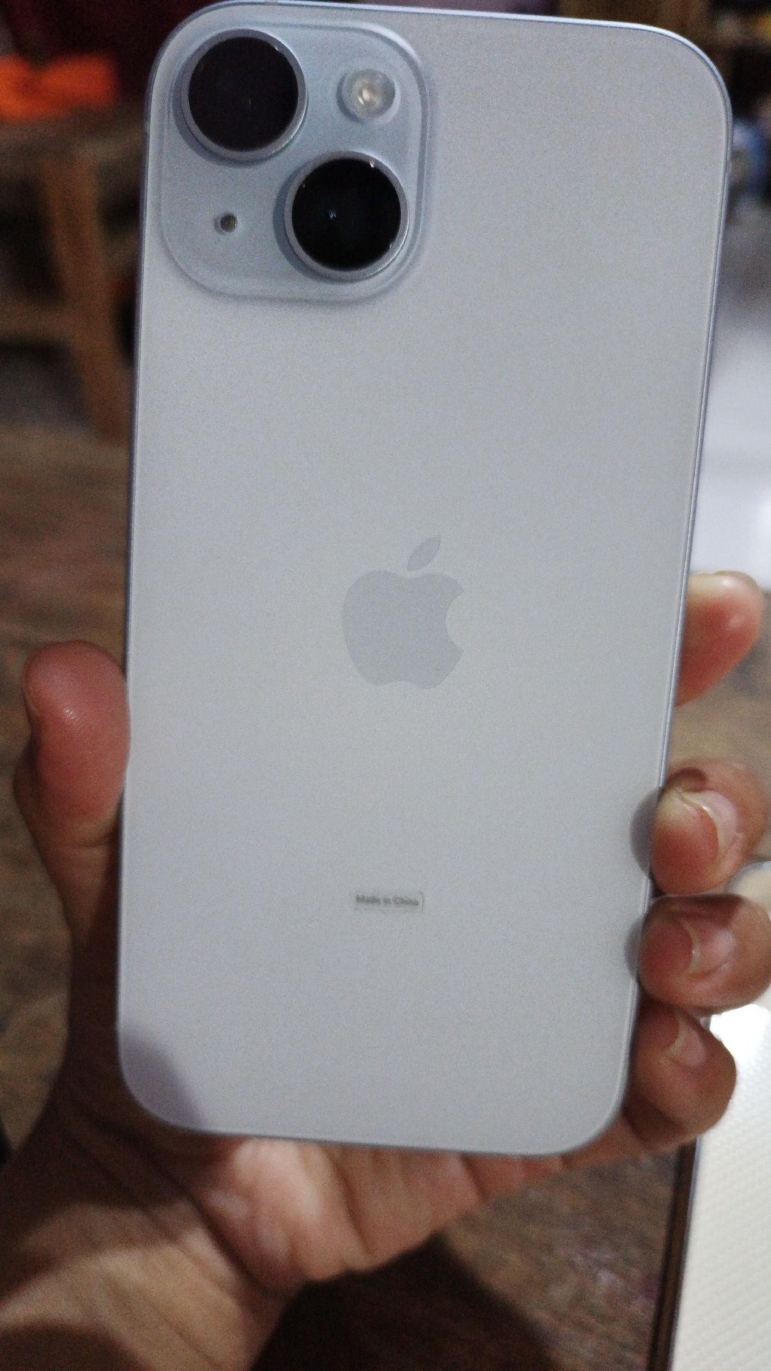 An overhead shot shows a light gray smartphone, likely an iPhone, being held by a hand. The back of the phone is visible, featuring a dual-camera system in the upper left corner, the Apple logo in the center, and "Made in China" text near the bottom. The phone is positioned vertically. The background appears to be a wooden surface and out-of-focus elements that could be furniture. The scene is indoors, possibly in a residential setting. The location is not specified in the image or its context provided.
