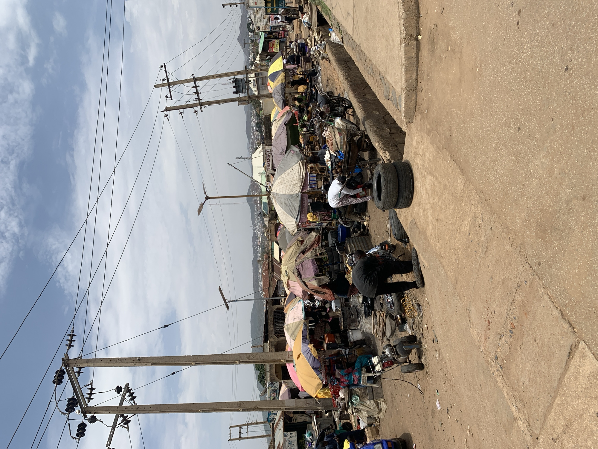 The image depicts a bustling street scene in Rafin Par, Nigeria. Overhead, power lines are strung between utility poles against a backdrop of a partly cloudy sky. The street is lined with vendors and their stalls, sheltered by a variety of umbrellas and awnings. People are engaged in various activities, including what appears to be tire repair. The scene is a mix of commercial activity and everyday life. The location is defined by the visual cues within the environment.