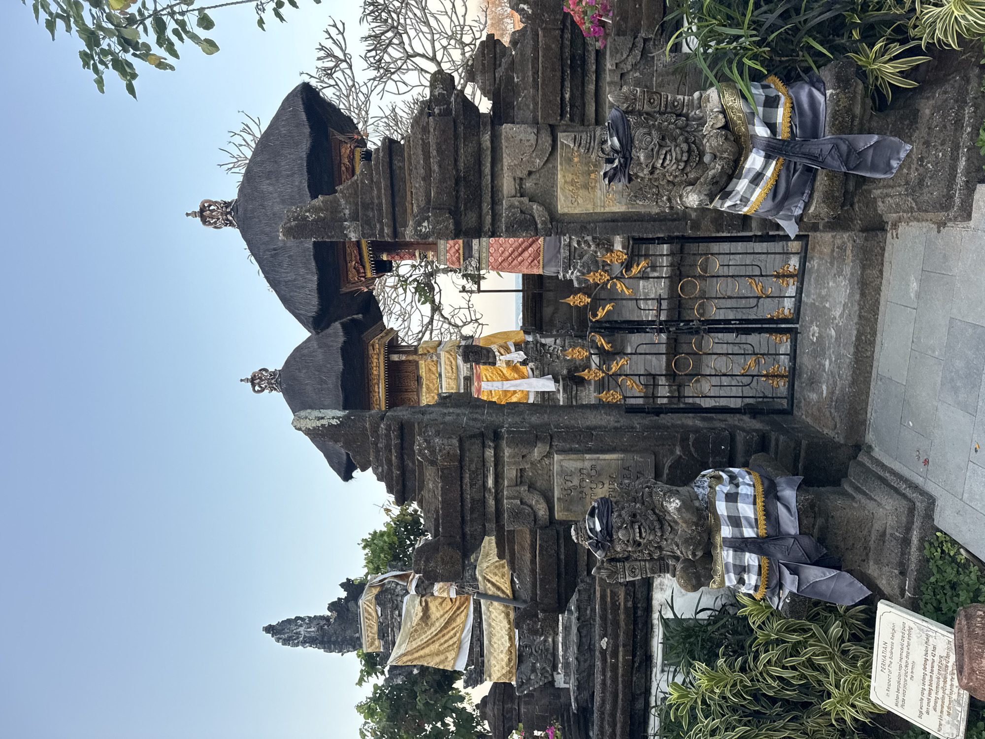 The image shows the entrance to a Balinese temple, likely Pura Luhur Uluwatu, located in Kuta Selatan, Indonesia. The foreground features two stone guardian statues, dressed in traditional Balinese checkered fabric. Behind them is an ornate black metal gate with gold accents. The gate leads to a stone staircase, flanked by layered stone structures adorned with yellow and white fabric. Two thatched-roof structures with intricate golden crowns are visible in the background. Lush green foliage and bare trees surround the temple. The sky is clear and pale blue. A sign in the lower left corner provides information about temple etiquette.