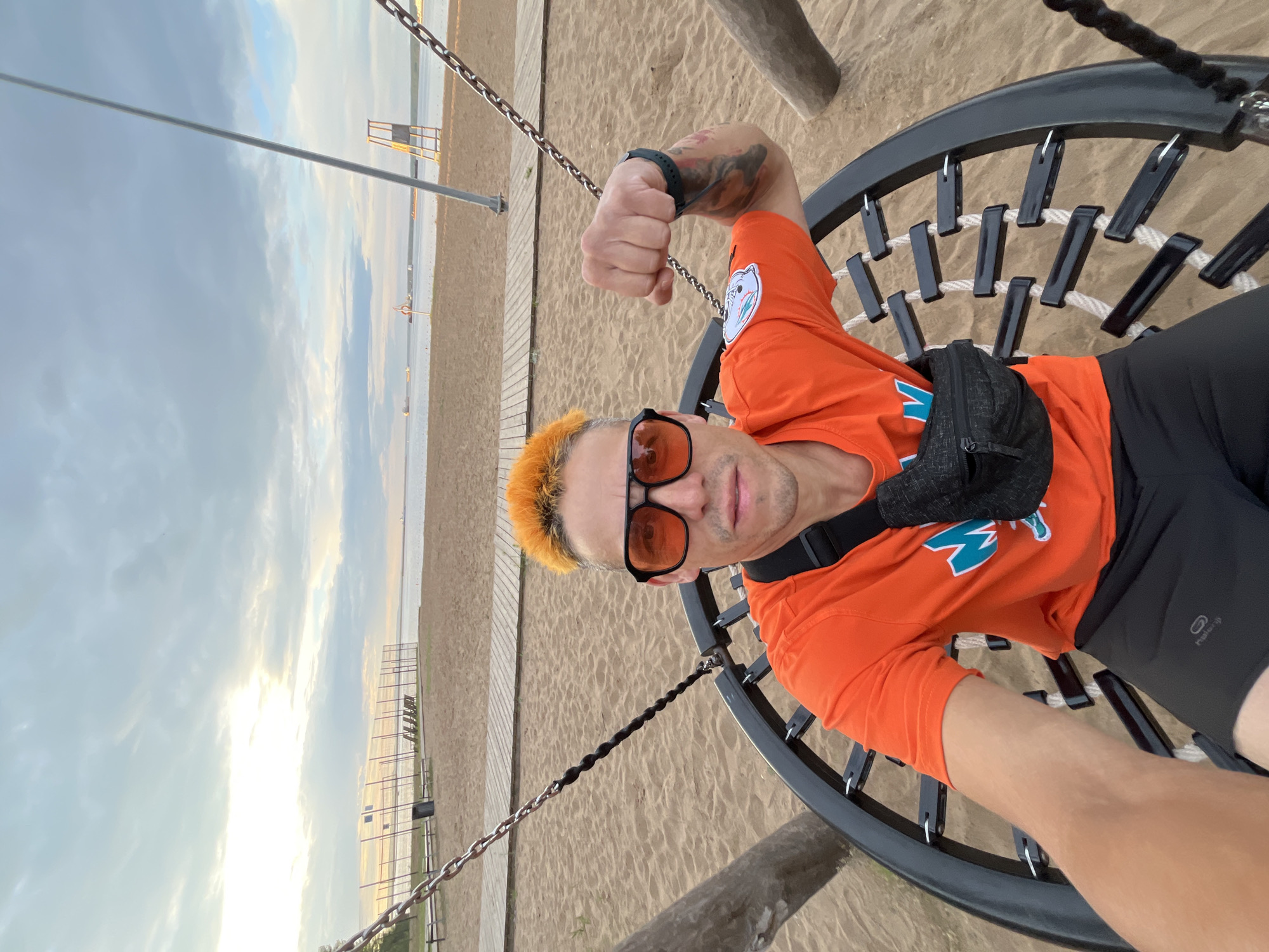 The image features a selfie of a man with bright orange hair, wearing an orange t-shirt and sunglasses. He is sitting in a circular swing with a dark frame and rope netting. The man has tattoos on his arm and a black fanny pack around his waist. He appears to be looking at the camera with a neutral expression.
The background shows a sandy beach with wooden boardwalks and a body of water, likely a lake or sea, in the distance. There are also metal structures on the beach, possibly part of a playground or sports area. The sky is cloudy with hints of sunlight breaking through, suggesting it is either early morning or late afternoon. The overall scene conveys a relaxed, outdoor atmosphere, possibly a park or recreational area by the water. The location context is Chaikovsky, Russia. There is no visible text in the image.