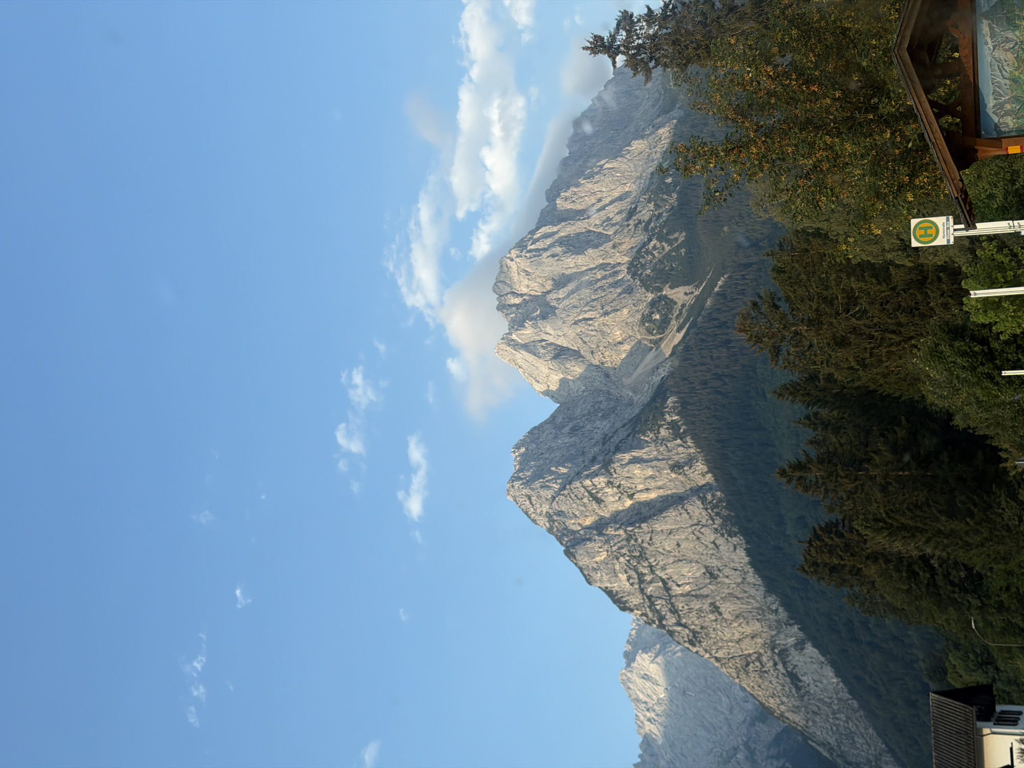 In Grainau, Germany, a dramatic mountain range dominates the scene under a clear blue sky with scattered white clouds. The rugged, rocky peaks are illuminated by sunlight, casting shadows that emphasize their textured surfaces. A dense forest of evergreen trees covers the lower slopes of the mountains, creating a stark contrast with the bare rock above. In the foreground, a wooden bus stop shelter with a visible map of the area is present, along with a green and yellow bus stop sign. The roof of a building is visible on the far left. The scene captures a serene and majestic natural landscape.