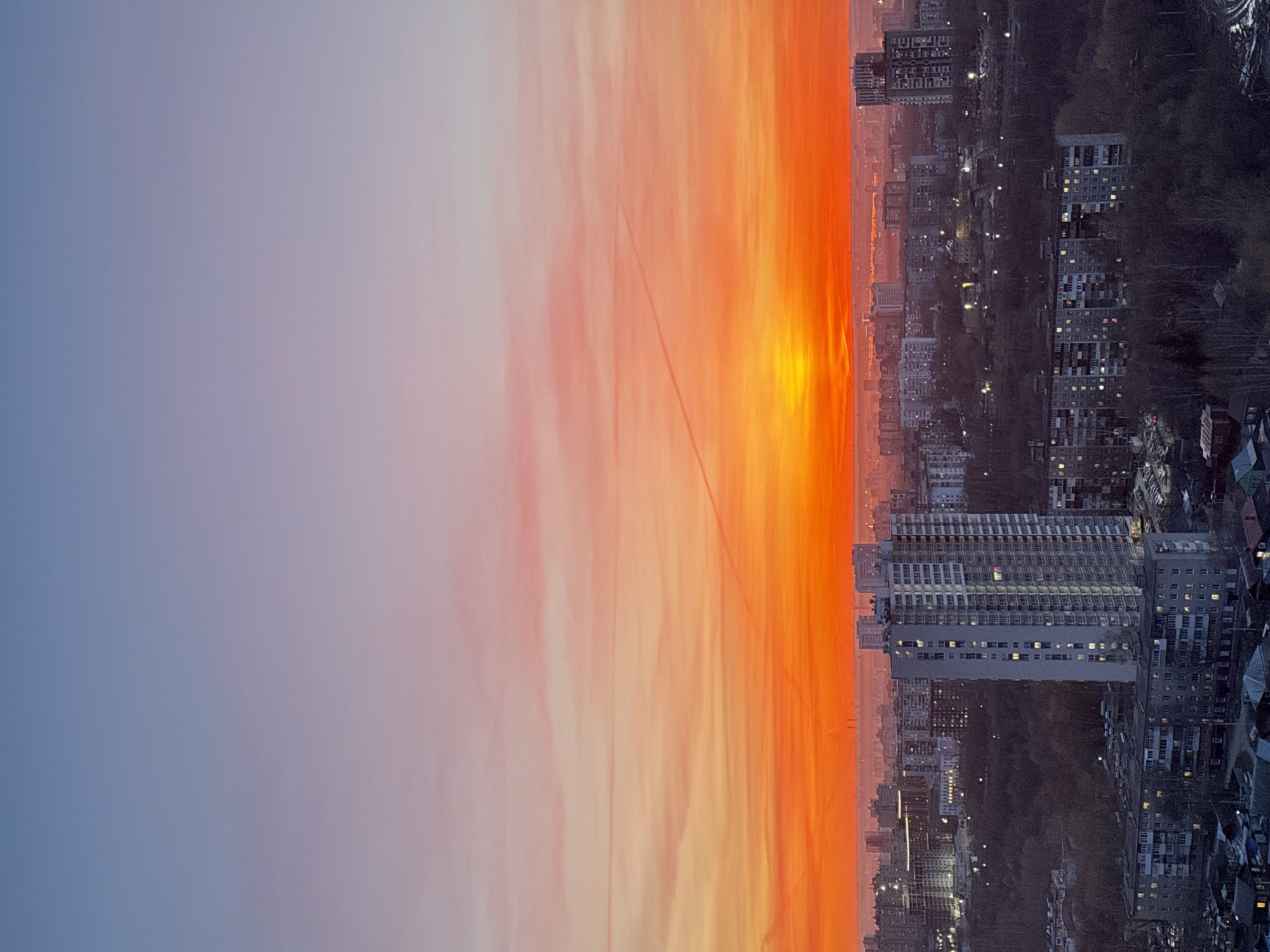 This panoramic image captures a breathtaking view of the city of Perm, Russia, during either sunrise or sunset. The sky dominates the upper portion, displaying a spectacular gradient of colors. At the very top, the sky is a soft, muted blue-grey, transitioning downwards through gentle purples and pinks. Closer to the horizon, the colors intensify dramatically into vibrant oranges and brilliant yellows, indicating the sun is just below or rising above the horizon. Streaky, elongated clouds catch these warm hues, adding texture to the expansive sky. A faint, thin line, possibly a power line or contrail, can be seen traversing the colorful expanse.
Below this radiant sky lies the sprawling cityscape of Perm. Numerous buildings, primarily multi-story residential and commercial structures, stretch across the landscape. Their forms are silhouetted against the bright horizon, appearing darker in contrast to the illuminated sky. Many windows within these buildings are lit up, creating a delicate pattern of twinkling lights that suggests the city is active as day transitions to night, or vice versa. The urban area appears dense in some parts, with hints of darker, possibly wooded or undeveloped patches interspersed throughout. The overall impression is one of a vast, active city under a dramatically painted sky, evoking a sense of tranquility and awe at the natural display of light. There is no visible text in the image.