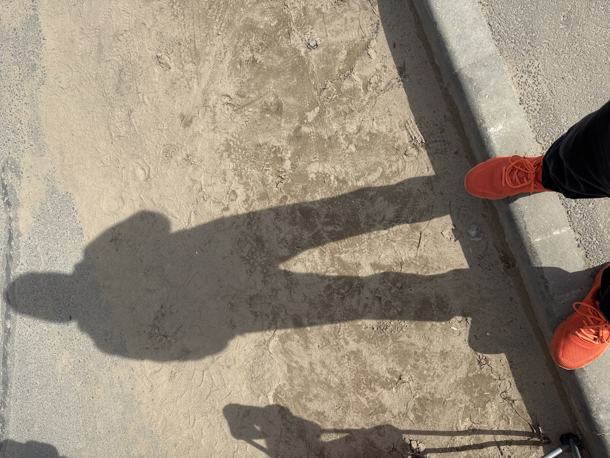 The scene depicts an outdoor location in Kaduy, Russia. The ground consists of packed dirt with numerous footprints. A shadow of a person is cast onto the dirt. The person is wearing dark pants and orange athletic shoes, and is partially visible in the upper right and lower right corners. Part of a metal tripod is visible in the lower right corner. To the right of the dirt ground is a section of concrete curb. To the left of the dirt ground is an area of paved road.