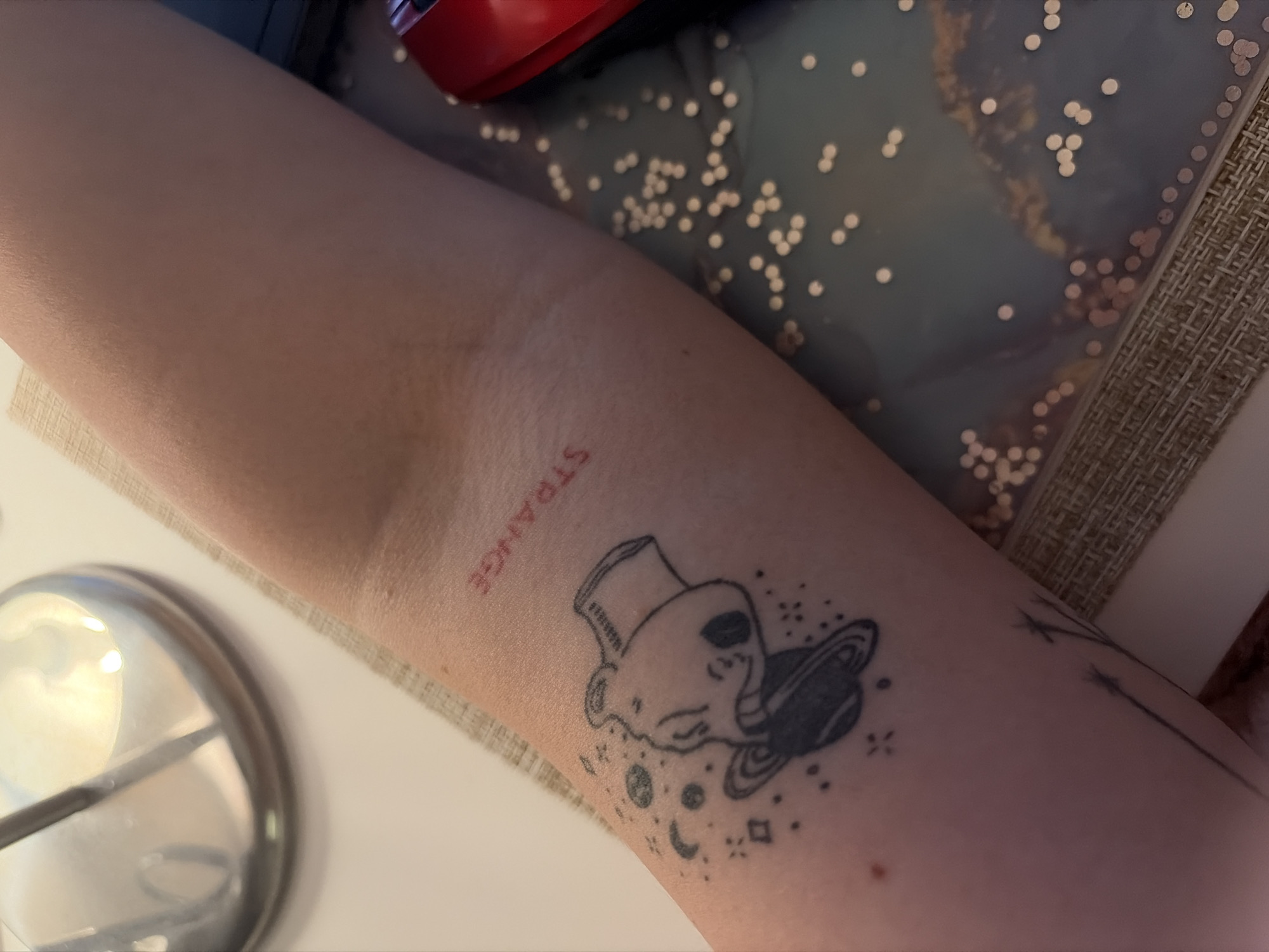 An arm rests on a surface. There is a tattoo of an alien with a planet on the arm. The word "strange" is tattooed above the alien. There is a lamp next to the arm. The image was not taken in Vozdovac Urban Municipality, Serbia.