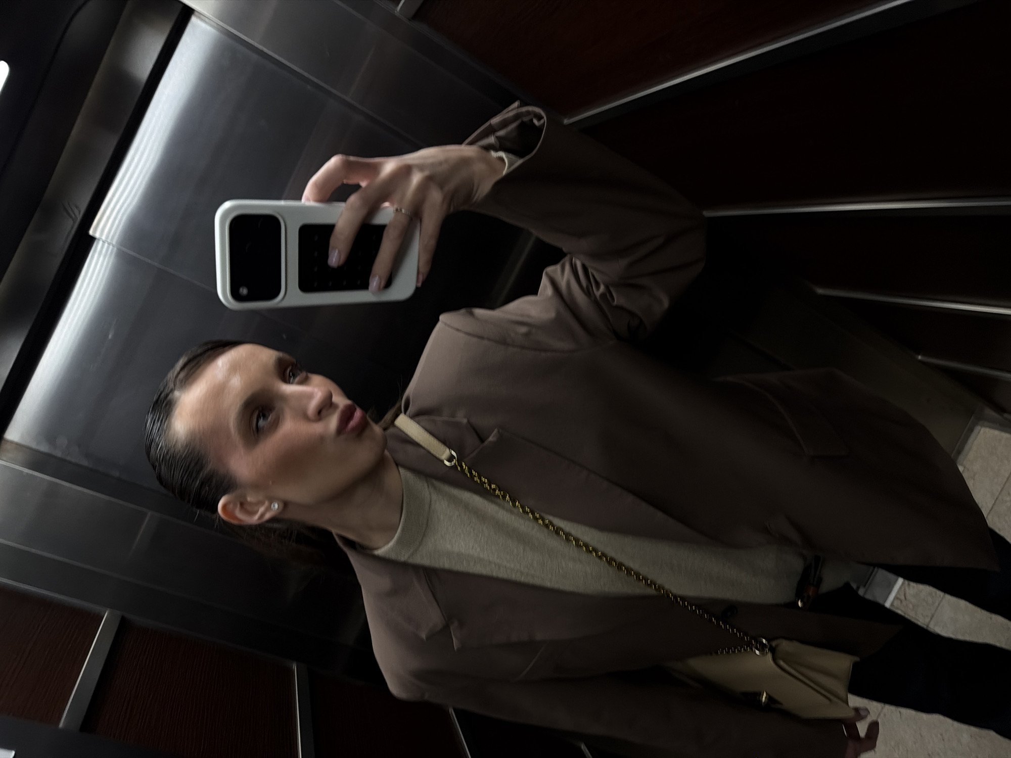 A woman takes a selfie in an elevator. She holds a smartphone with a white case in her right hand. She has dark hair pulled back, wears a light tan sweater under a brown blazer, and a small beige purse with a gold chain. The elevator walls are silver and dark brown, reflecting the scene. The location is unknown, and no information suggests it's in Budva, Montenegro.