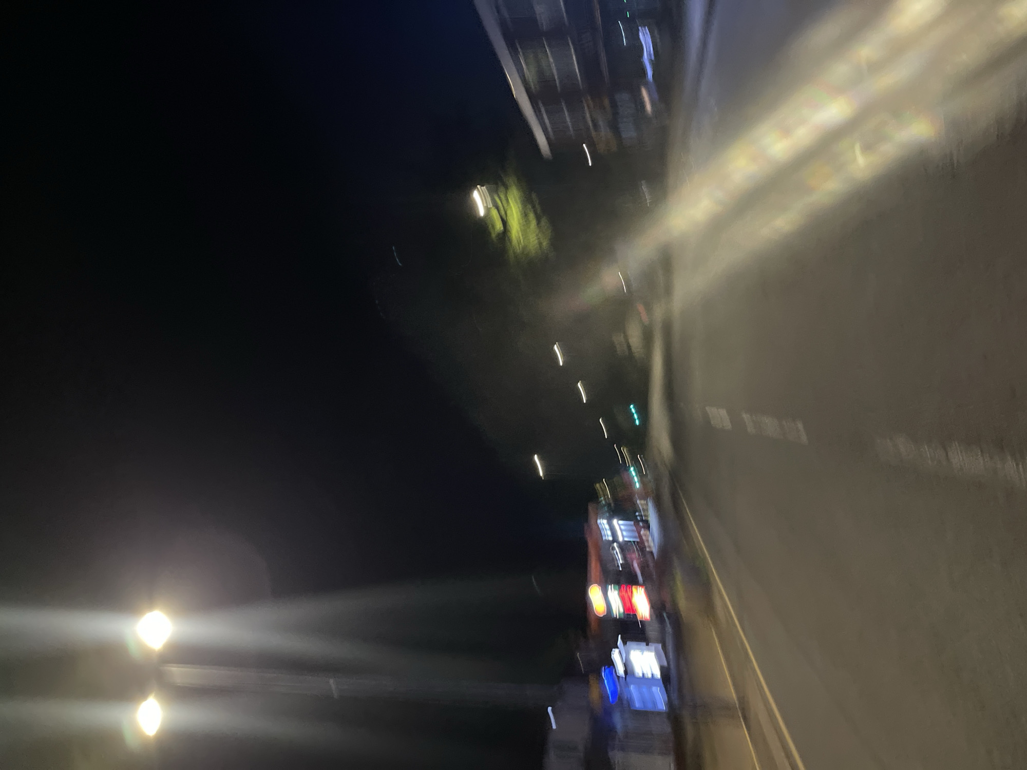The image depicts a night scene in Maidstone, United Kingdom. A street with a wet, reflective surface stretches into the distance. Streetlights are visible along the road and on buildings. The buildings show various illuminated signs, suggesting commercial activity. A portion of the scene is in darkness on the left, with a few illuminated streetlights. The image exhibits motion blur, possibly from a moving vehicle or handheld camera.