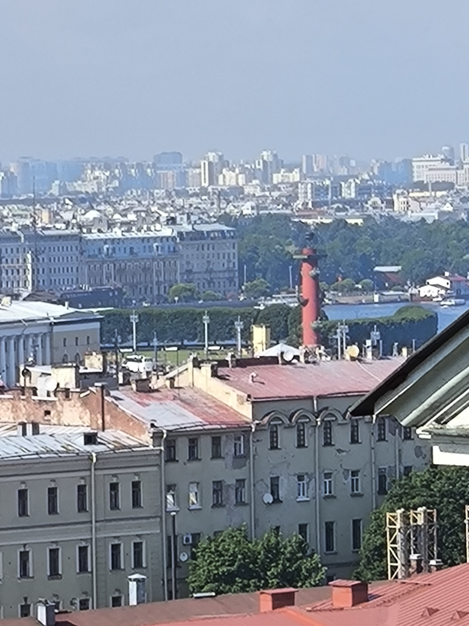 The image shows a panoramic view of the cityscape of Saint Petersburg, Russia, on a clear day. In the foreground, several buildings with red-tiled roofs are visible, some showing signs of wear and tear. Behind these, more buildings with grey and light-colored facades and numerous windows can be seen.
Dominating the middle ground is a prominent red lighthouse-like tower, situated on a body of water. The tower appears cylindrical and has a conical top. Green trees and manicured hedges surround the base of the tower and line the waterfront. In the distance, a sprawling urban landscape with numerous high-rise buildings stretches to the horizon under a pale sky. The overall scene depicts an urban environment with a mix of historical and modern architecture. There are no people visible in the image.