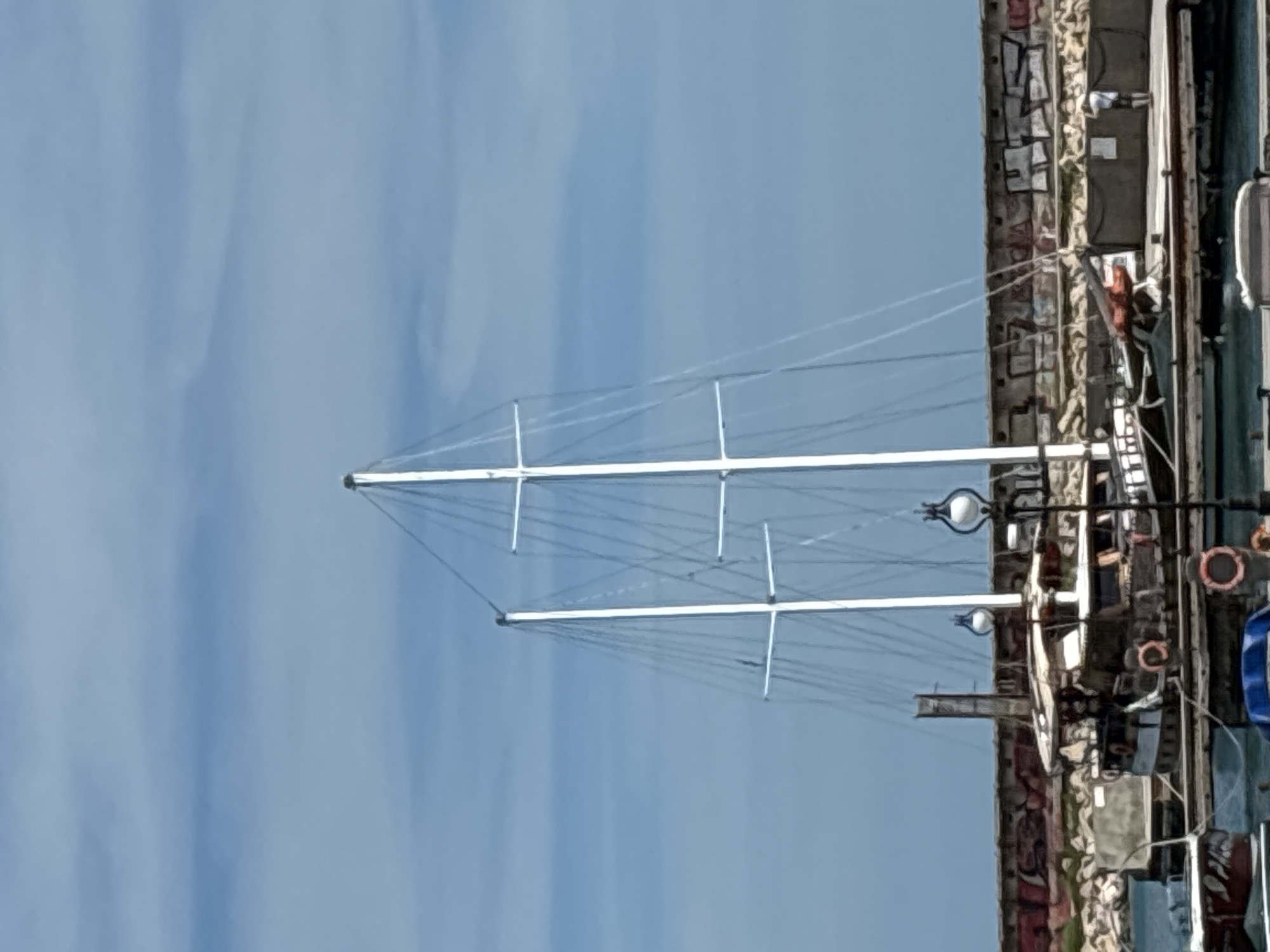 A medium shot shows two masts of a sailboat anchored at a harbor in Varna, Bulgaria. The boat has a dark hull with white trim and several life preservers. The masts are tall and white, with rigging. In the background, a stone wall with graffiti runs along the harbor, and beyond that, a person is standing on a dock. The sky is blue with scattered clouds.