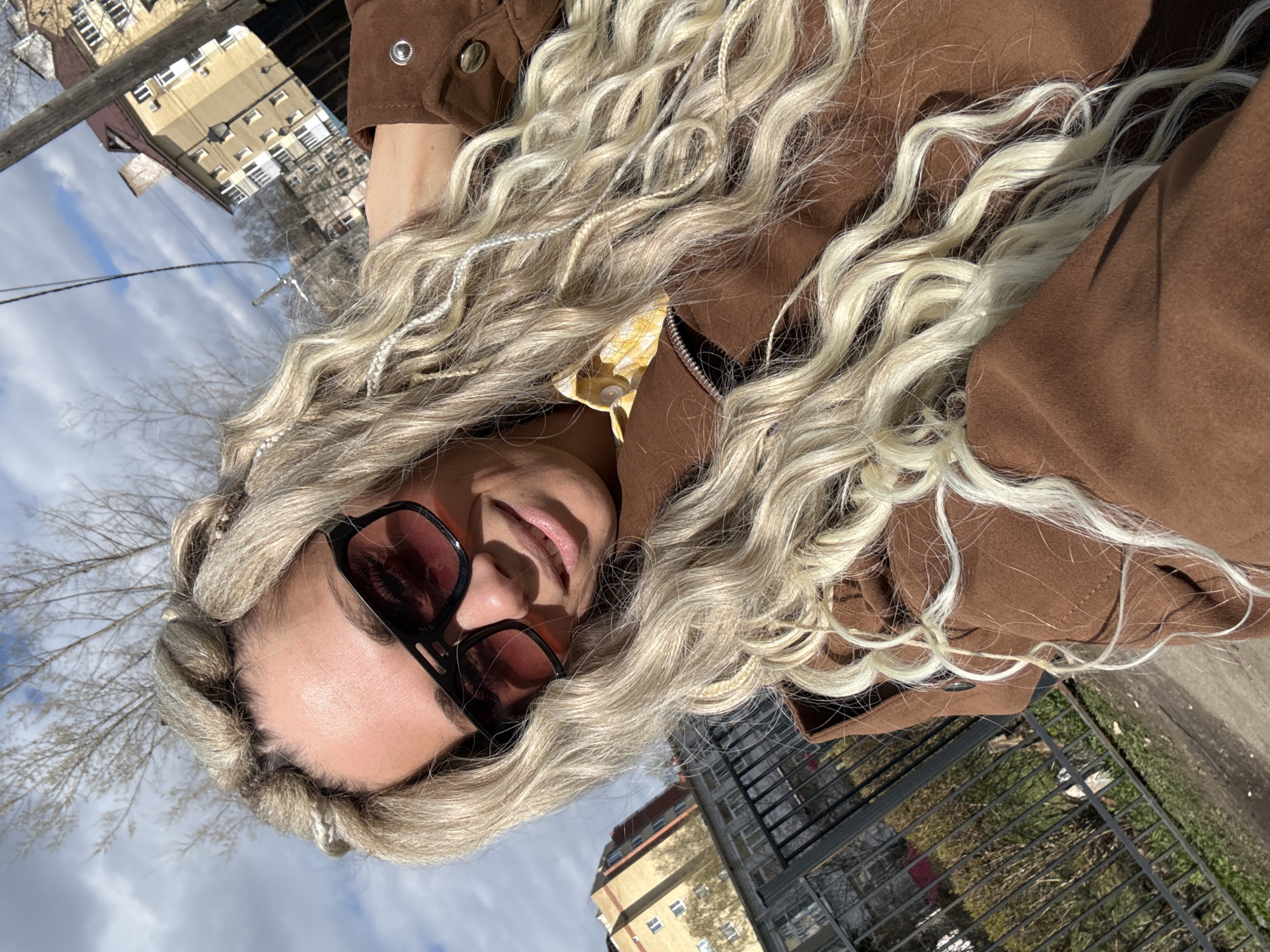 A woman with long, blonde braided hair is wearing sunglasses and a brown jacket. She is outdoors in what appears to be an urban environment. In the background are buildings, possibly apartment complexes, and a cloudy sky. The location could be Rusanovo, Russia, based on the prompt. There are bare trees and what looks like a fence or railing in the background.