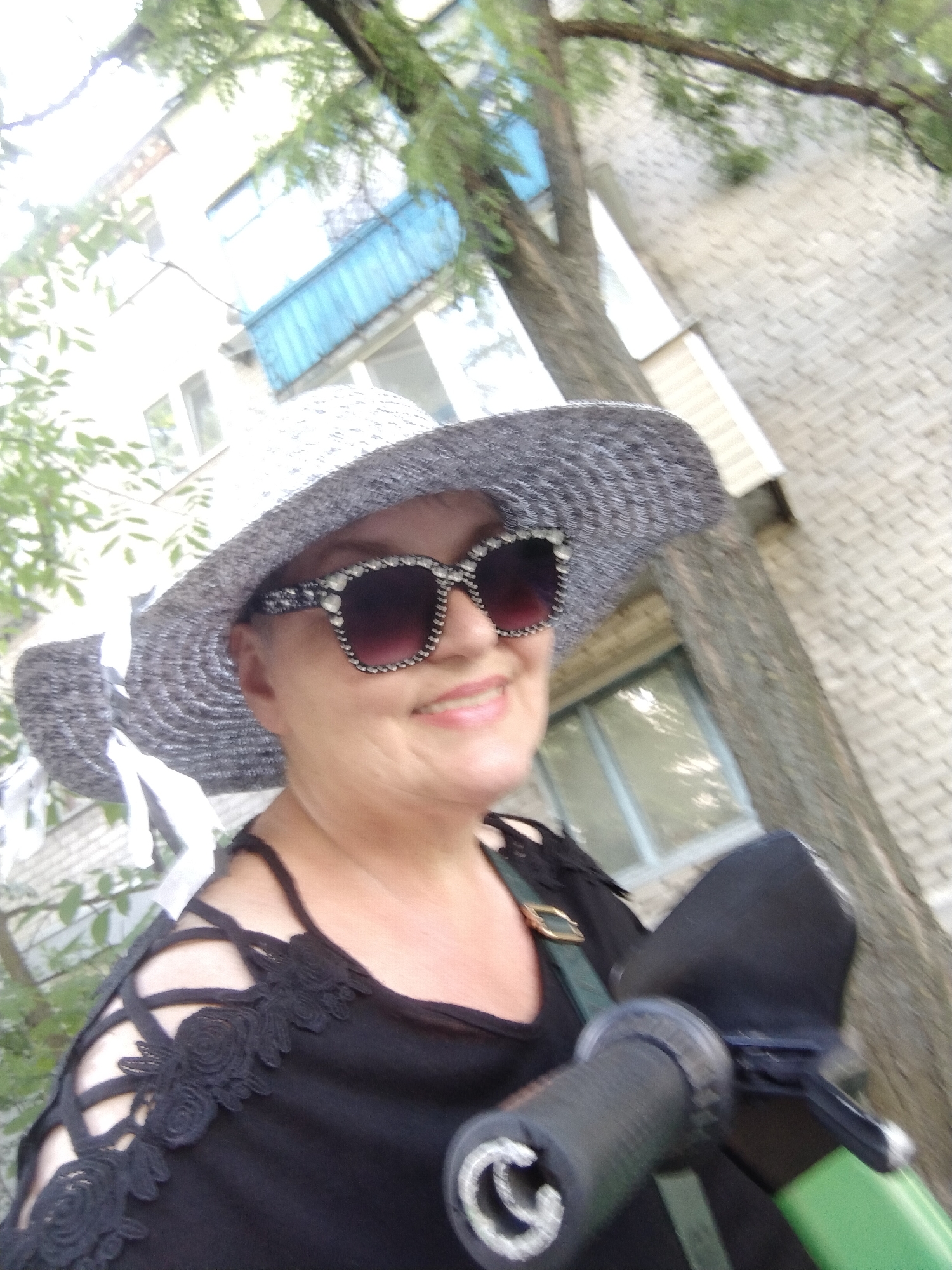 A woman is pictured outdoors, in what appears to be an urban setting in Horishni Plavni, Ukraine. She is wearing a wide-brimmed hat adorned with white ribbons, sunglasses with decorative rhinestones, and a black top with intricate lace detailing on the shoulders and straps. The background shows a brick building with windows, trees with green foliage, and a blue balcony. The woman is holding onto the handlebars of a green scooter, suggesting she may be on the move or about to depart. The overall scene conveys a sense of casual outdoor activity.