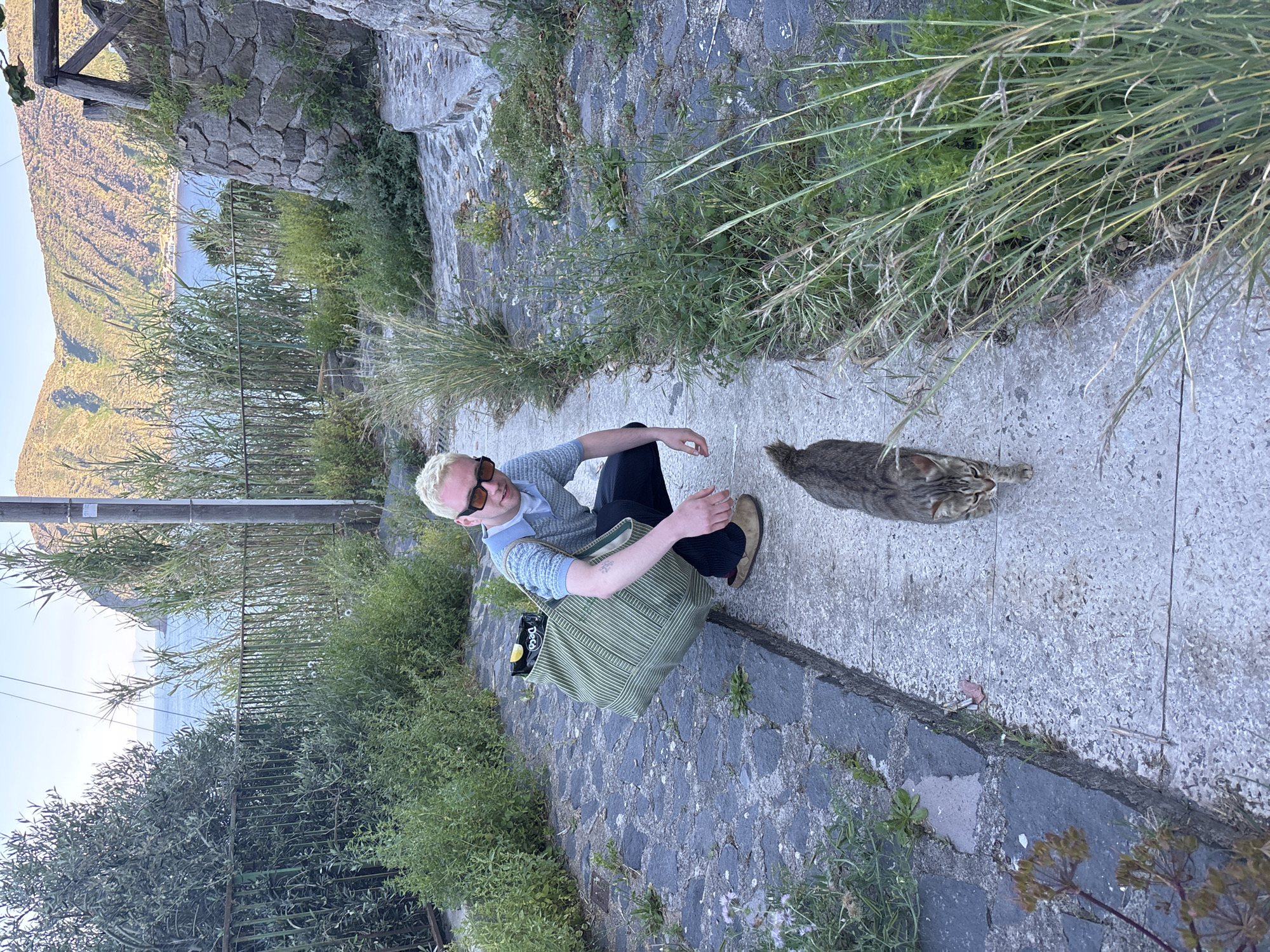 A man is sitting on a concrete pathway in Lipari, Italy, next to a tabby cat. The man has short, bleached blond hair and is wearing sunglasses, a light blue sweater, and dark pants. He is also holding a green striped tote bag. The cat is lying down on the pathway, looking towards the man. The pathway is surrounded by vegetation and a metal fence. In the background, there is a view of the sea and mountains. The sky is clear. The path appears to be on the side of a building or structure with visible stonework.