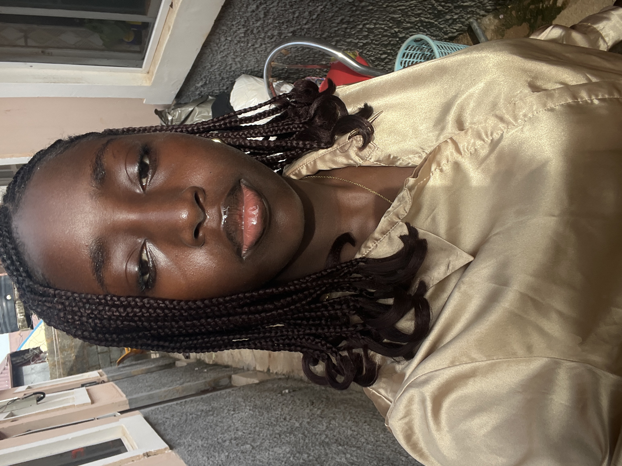 A medium shot of a young woman with dark skin, braided hair, and glossy lips is angled upward. She is wearing a gold-colored satin top, with part of a thin gold necklace visible. The background is indistinct, appearing to show an exterior location. Some concrete surfaces are visible along with what looks like a window frame, and other blurred objects. Specific details to confirm the location as Abuja, Nigeria are not evident in the provided image.