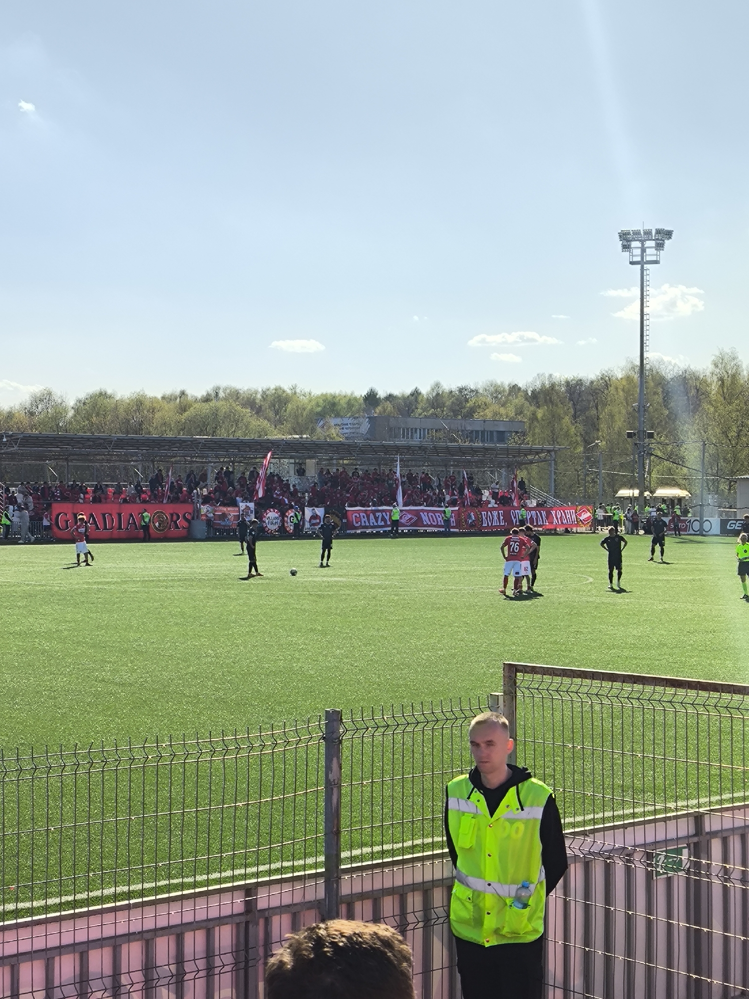 The image depicts a soccer field with a game in progress in Moscow, Russia. A crowd is gathered in the stands behind a large "GLADIATORS" banner, also displaying other banners in Cyrillic. Several players are on the field, some in red and others in black. A man wearing a bright yellow vest stands in the foreground, near a fence. A tall metal light tower is visible in the background, along with trees and a bright blue sky.