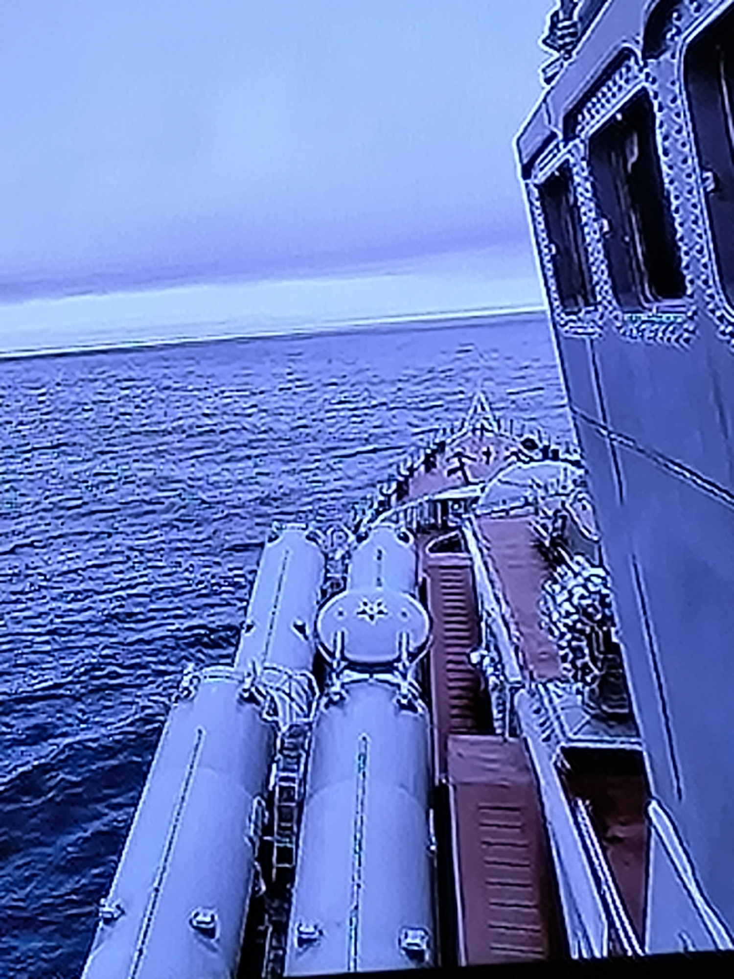 The image shows a view from a ship at sea. The water is dark blue with small waves. In the foreground, there are several large, cylindrical objects, possibly missile silos, lined up on the deck of the ship. The ship itself appears to be a warship, with a large superstructure visible on the right side of the image. The sky is a pale blue, with some clouds on the horizon. The image does not provide any information that would indicate it was taken in the city of Kazan, Russia.