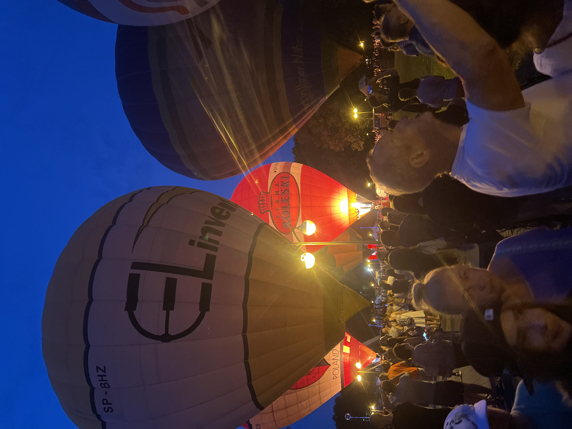 The image depicts a hot air balloon festival at dusk in the city of Tychy, Poland. In the foreground, a crowd of people is gathered, some with their arms raised, possibly taking photos or videos with their phones. The hot air balloons are the central focus, with several inflated and illuminated, casting a warm glow on the scene. One balloon in the foreground has the text "EL Invest" and "SP - BHZ" visible. Another balloon, predominantly red and white, displays "FIRMA KOLESKI" and "-SP-BH". The background shows trees and lights from the festival grounds. The sky is a deep blue, indicating it is twilight. The overall atmosphere appears festive and lively.