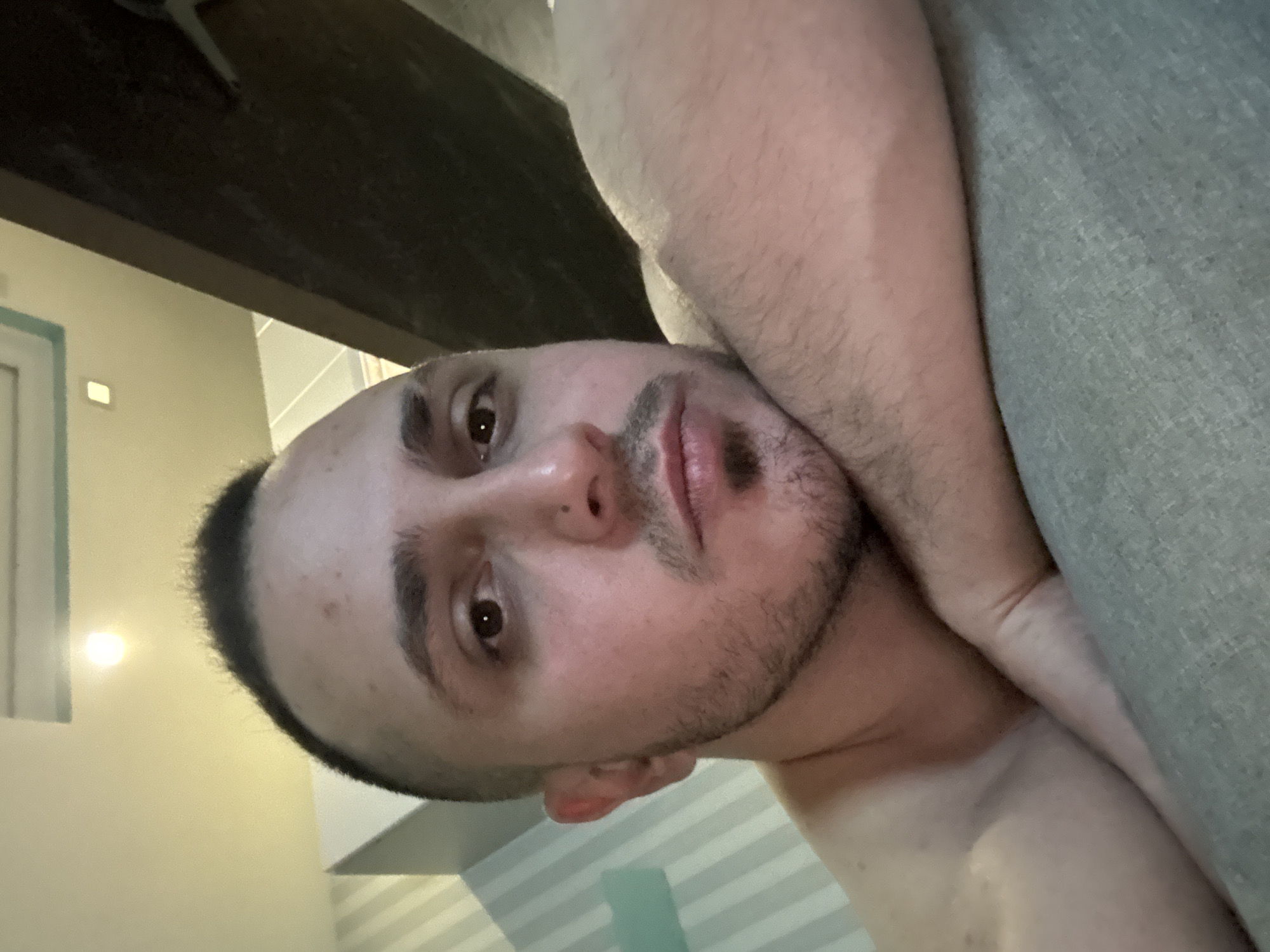 The media file displays an indoor portrait, rotated 90 degrees clockwise from its natural orientation. The primary subject is a male-presenting individual, seen in a close-up, likely reclining or lying down. He has short, dark hair (a buzz cut), light skin, and stubble on his chin and upper lip. His gaze is directed towards the camera, with a neutral to slightly contemplative expression.
The person's bare upper torso and arm are visible, with one arm appearing to rest on or next to a grey, textured fabric, possibly a blanket, pillow, or upholstery of a couch/bed. In the background, there's a dark, horizontal structure, possibly a beam or shelf, across the top. To the left, a light-colored wall or doorframe is visible, featuring a bright light source (likely a light fixture) on it. Below the individual's head, patterned wallpaper or a striped surface can be seen, adding depth to the indoor setting. The overall lighting is somewhat dim, highlighting the direct light source. No text is visible in the image.