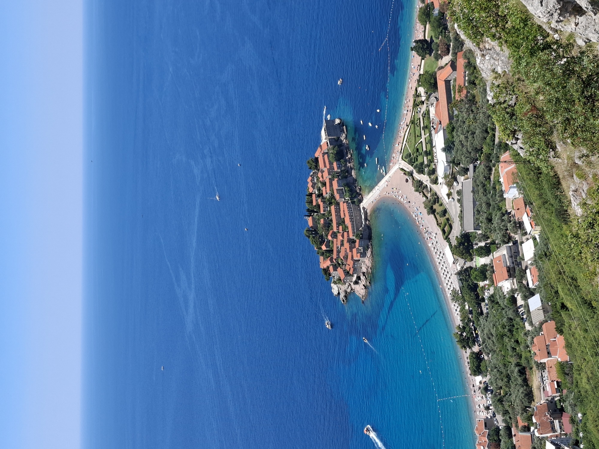 This is a high-angle, wide shot of the Sveti Stefan island resort in Montenegro, a luxury hotel complex located on a fortified island. The island is connected to the mainland by a narrow tombolo, a sandbar. The buildings on the island have traditional red-tiled roofs and are situated among lush greenery. The waters surrounding the island are a vibrant turquoise, with several boats visible. A sandy beach with numerous umbrellas and sunbathers is located on the tombolo. The foreground of the image shows a hillside covered with olive trees and more buildings with red-tiled roofs, suggesting a residential area. The sky is clear and blue, indicating a sunny day.