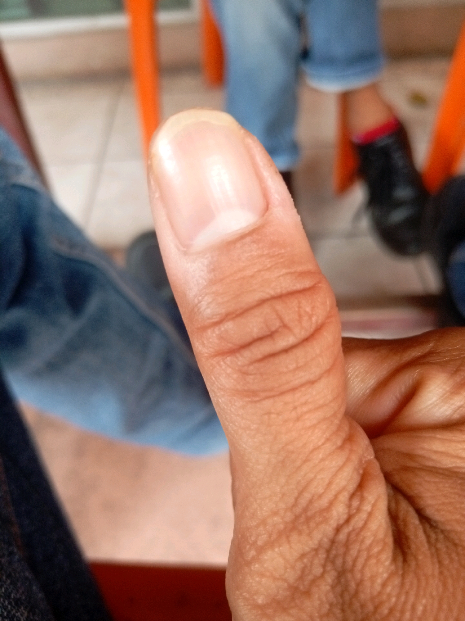 A close-up, slightly angled shot focuses on a person's thumb, showing the texture of the skin and the nail. The background is blurred but suggests an outdoor setting with orange chairs and people wearing denim jeans and black shoes. No specific location in Batam, Indonesia, can be determined from the image.