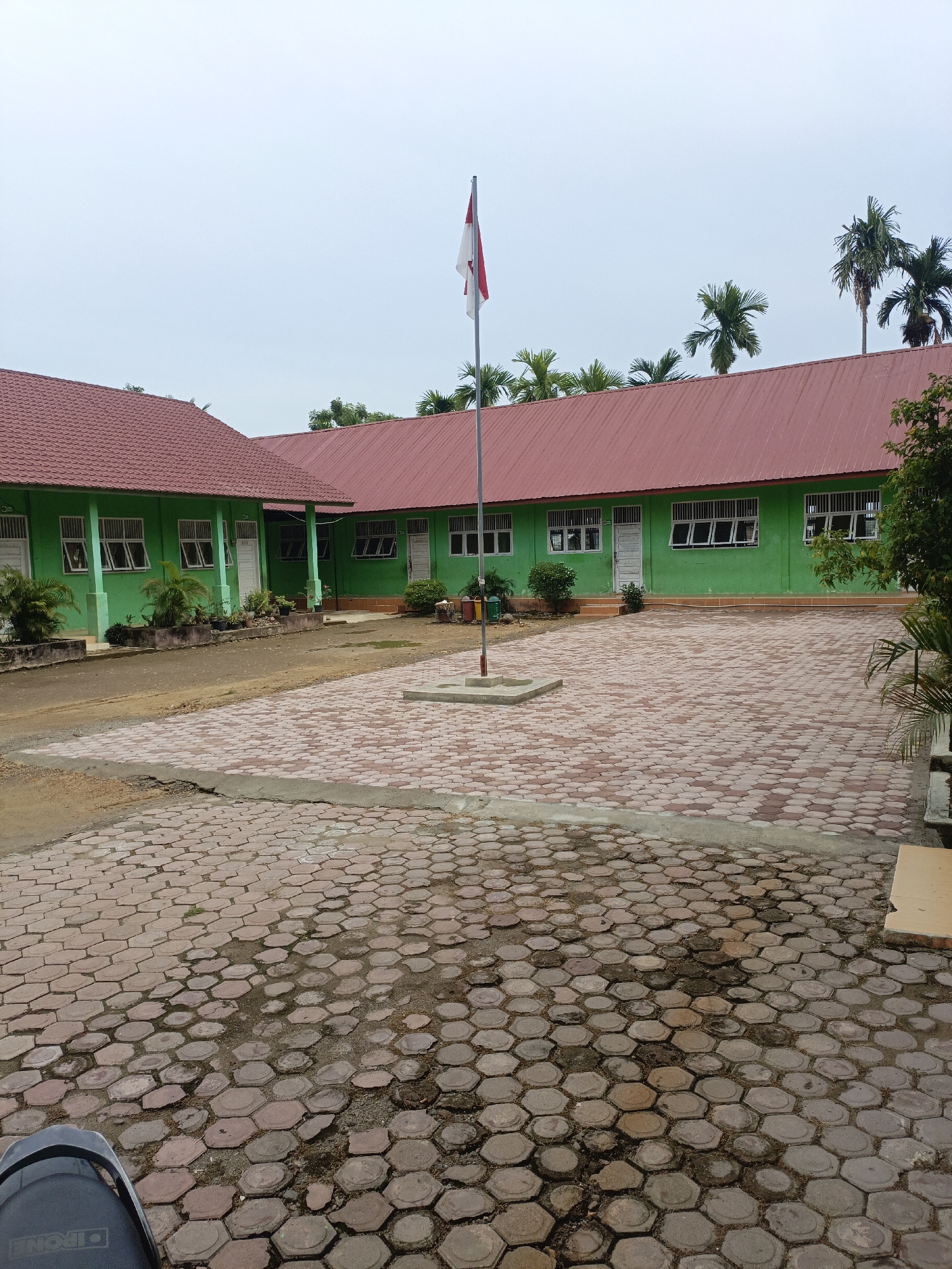 The image depicts a school building with a red-tiled roof and green walls, characteristic of many educational institutions in Lhokseumawe, Indonesia. The building appears to be a single-story structure with multiple windows and doors. In front of the building, there is a paved courtyard made of hexagonal concrete blocks. A flagpole with the Indonesian flag is prominently displayed in the center of the courtyard. Palm trees and other foliage are visible in the background, suggesting a tropical environment. The sky is overcast, indicating a cloudy day. The scene appears to be devoid of people, giving a sense of quietness and emptiness to the school grounds. The overall impression is that of a typical Indonesian school on a non-operational day, possibly during a holiday or the weekend. The presence of the Indonesian flag suggests this is a public building, possibly a government-run school.