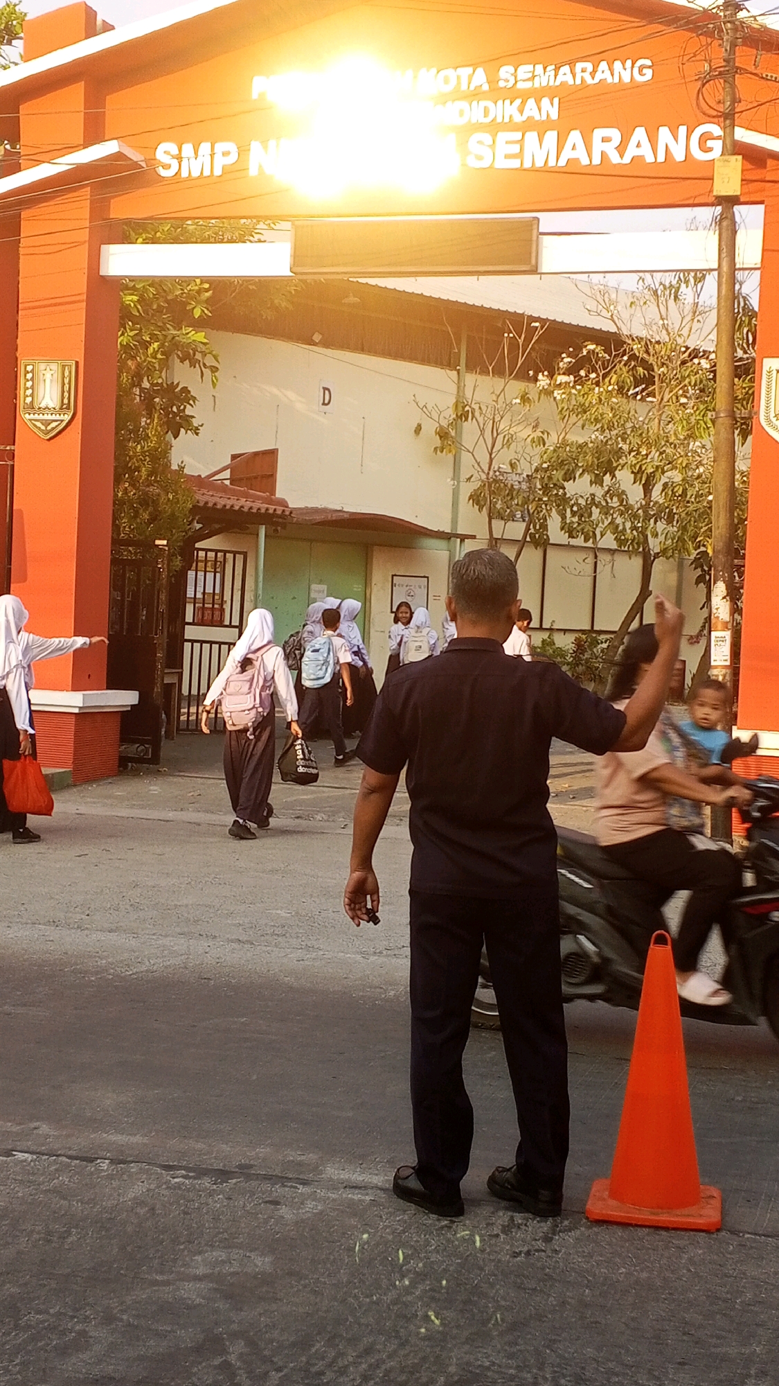 The image depicts the entrance of SMP Negeri 16 Semarang, a junior high school in Semarang, Indonesia. The school's orange gate is prominent, with text indicating "PEMERINTAH KOTA SEMARANG DINDIKNAS SEMARANG" and "SMP N 16 SEMARANG". The sun is shining brightly overhead, causing lens flare. Several students in school uniforms, mostly wearing hijabs, are walking towards or away from the school. One student in a white hijab and black pants is pointing to the left. Another student in a white hijab and black pants is carrying a dark bag. A man in a dark blue uniform, possibly a security guard or teacher, stands in the foreground facing away from the camera, with his right arm raised. He is holding a small black object in his right hand. An orange traffic cone is placed on the asphalt road to his right. A motorcycle carrying two people is visible in the background on the right side of the image. The environment appears to be a school campus with a paved road and some trees.