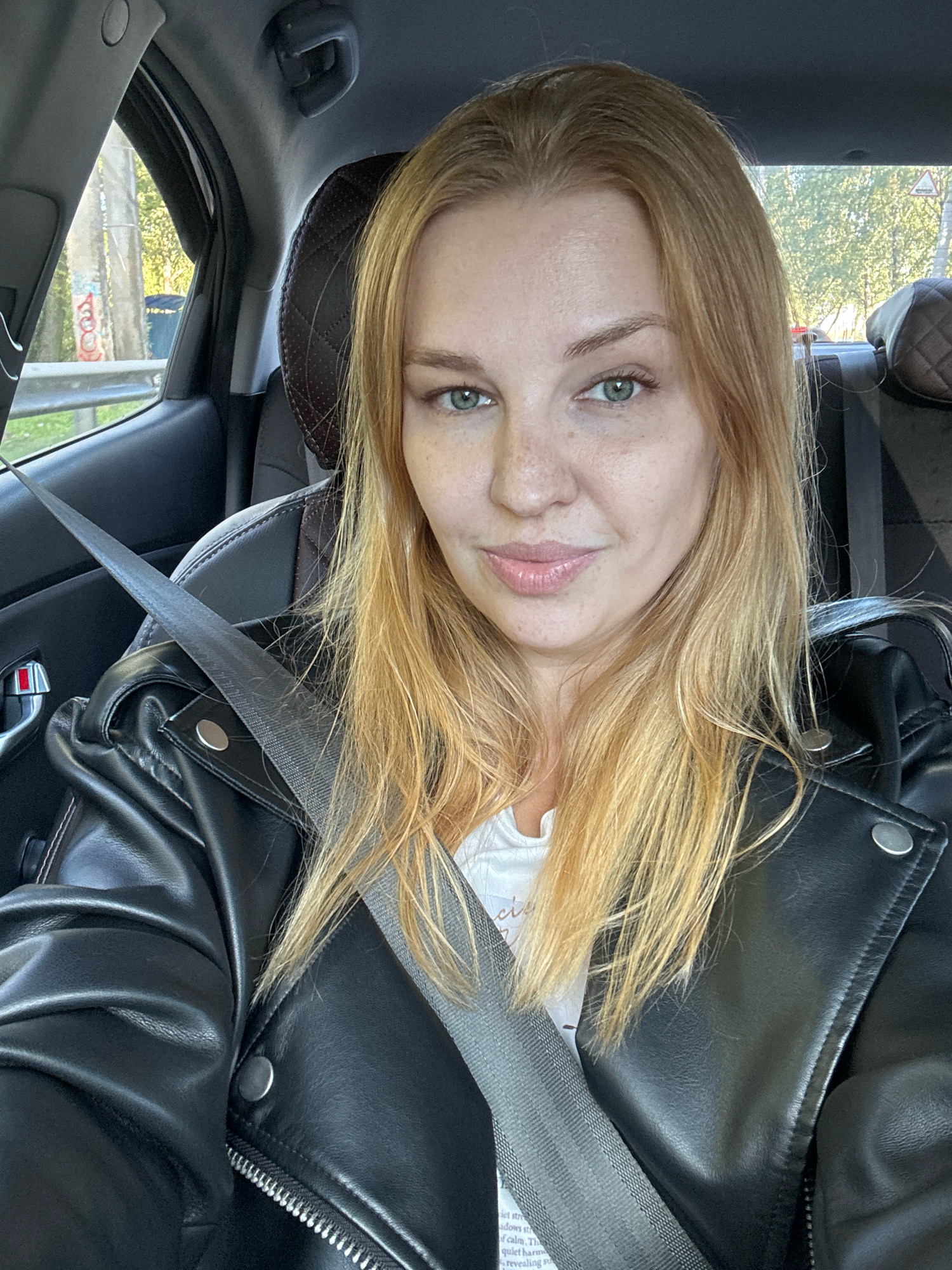A blonde woman with green eyes and freckles is taking a selfie while sitting in the passenger seat of a car. She is wearing a black leather jacket over a white t-shirt and a grey seatbelt is visible across her chest. The interior of the car is visible, including the headrest and door panel. Outside the car, trees and a road are visible. The scene is likely in Yaroslavl, Russia, as indicated by the signage visible in the background which appears to be Russian.