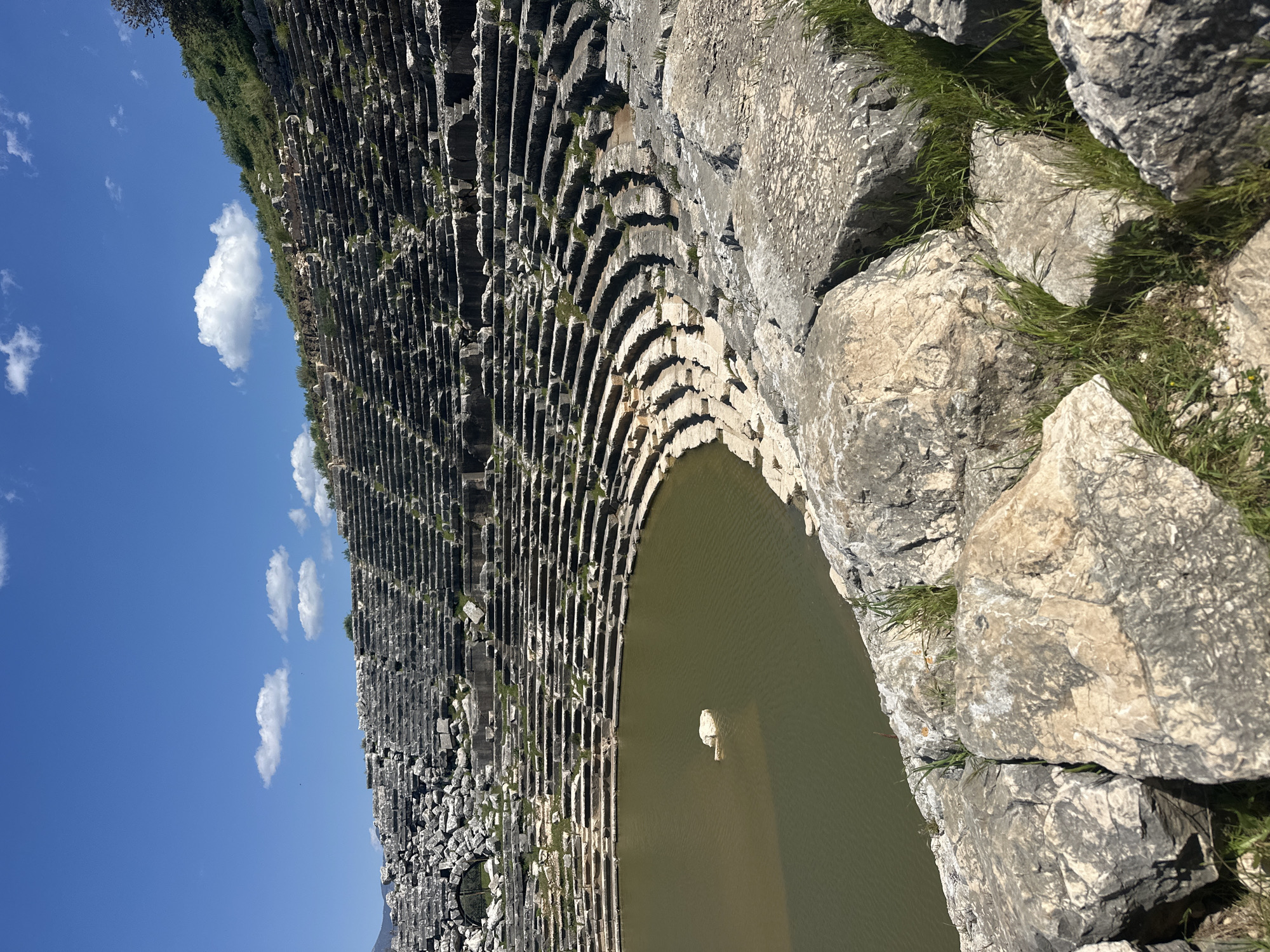 The image shows the ancient amphitheater in Tlos, located near the city of Seydikemer, Turkey. The amphitheater is partially filled with water, covering the stage area. Stone seating tiers rise from the water's edge. The structure is weathered and shows signs of age. Some stones are missing or displaced. A small section of sky with a few clouds is visible. Large rocks and some vegetation are present on the right side of the frame.