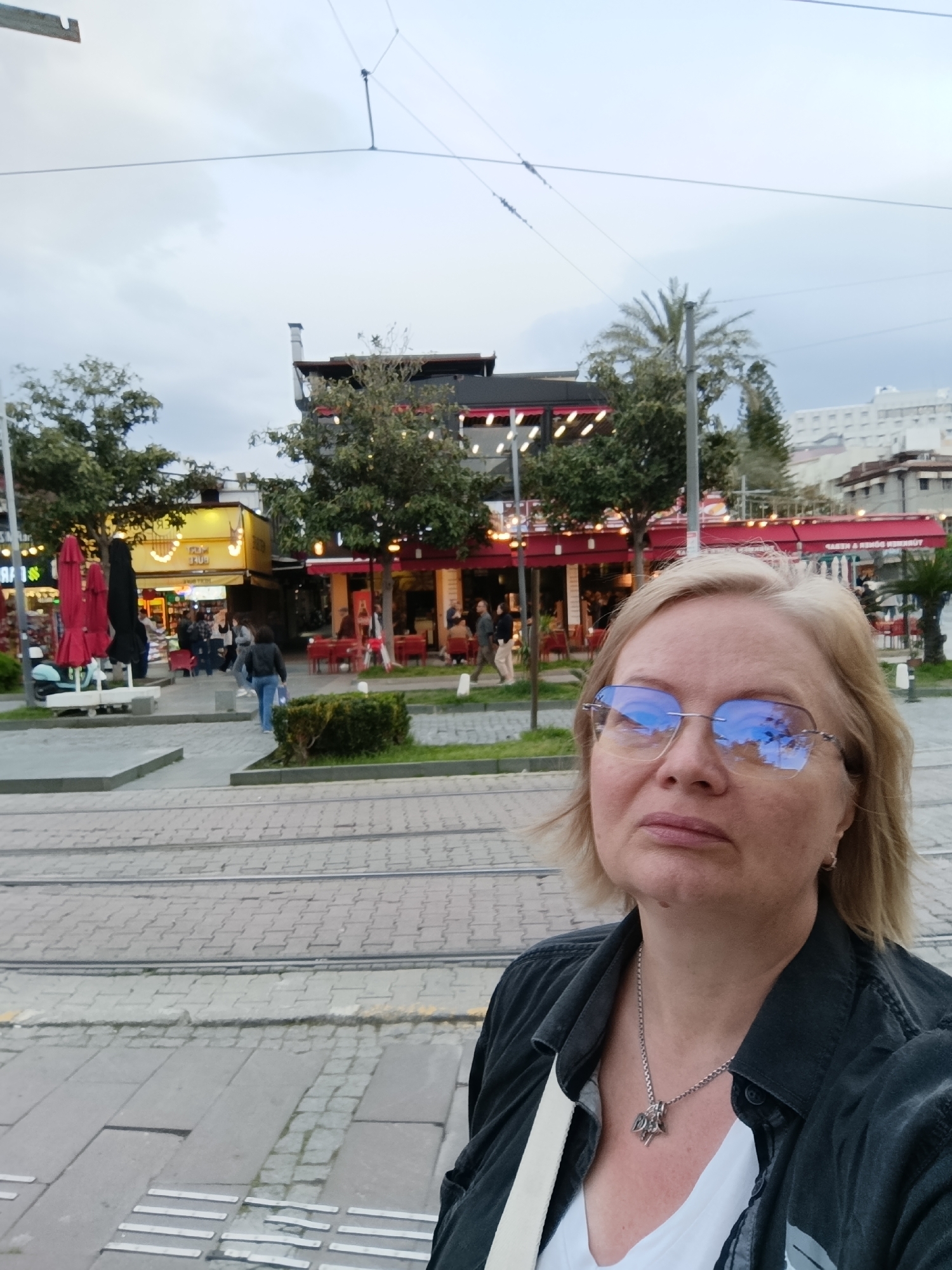 A woman takes a selfie in front of a public plaza. The woman has blond hair and wears glasses, a black jacket, a white top, and a silver necklace. She is standing on a light gray brick surface with tactile paving. Behind her is a pedestrian plaza with trees, shrubs, and various restaurants and shops. People are walking in the background. Overhead wires stretch across the scene. Based on architectural details, the location is likely in the city of Antalya, Turkey.
