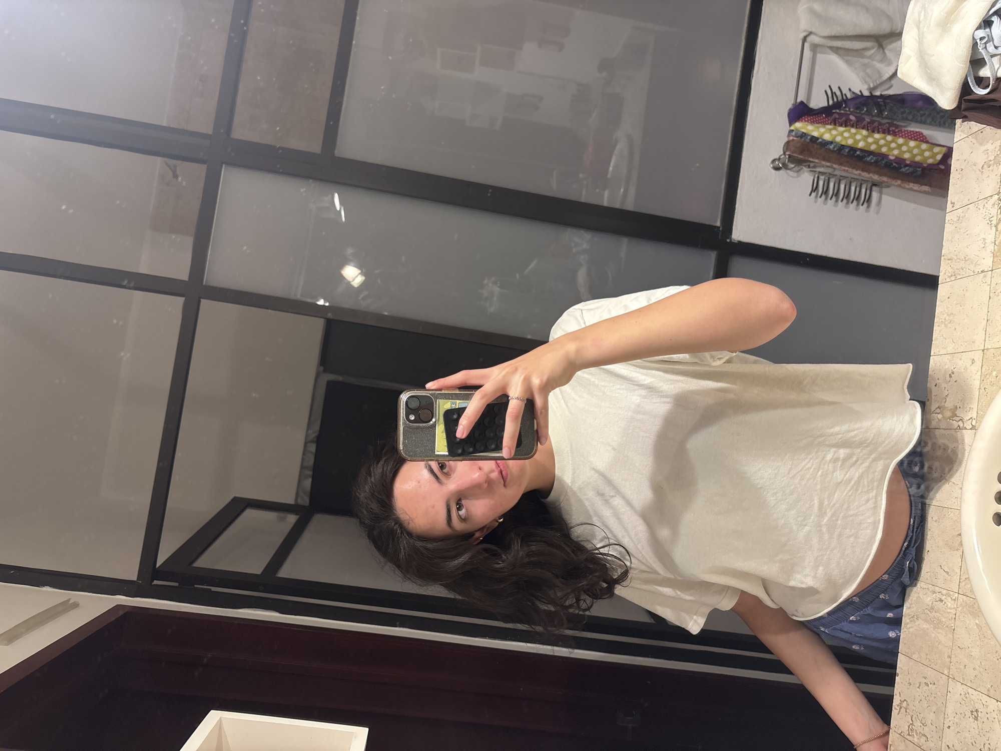 A person is taking a selfie in a bathroom. The person is wearing a white T-shirt and blue patterned shorts. The bathroom has a large mirror with black frames, a beige countertop, and a white sink. Behind the person, the mirror reflects the room, showing shelves and clothing items. The location is Puebla, Mexico.