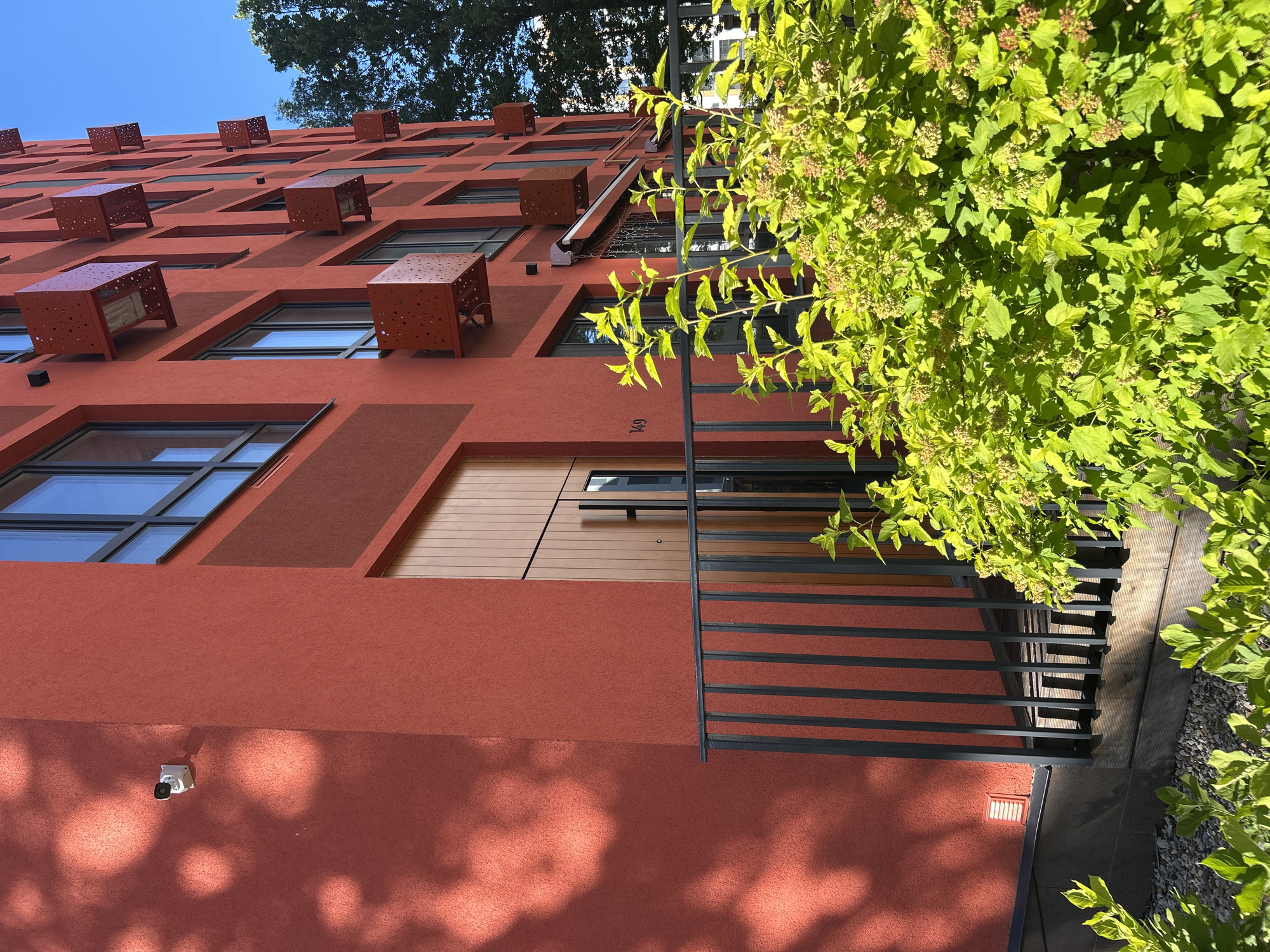 The media file displays a close-up, upward-facing view of a modern, red-walled building with numerous windows and distinctive, reddish-brown cube-like structures protruding from its facade. These structures, some with perforated designs, appear to be decorative or functional elements, possibly air conditioning units or unique architectural features. The building's texture is rough, suggesting stucco or a similar material.
Various windows of different sizes are visible, some reflecting a clear blue sky. On the lower right side of the frame, lush green foliage, likely from a tree or shrub, partially obscures the building, casting dappled shadows on the red wall. A black metal railing runs horizontally across the middle section of the building, hinting at balconies or a walkway. A security camera is mounted on the lower part of the wall.
The lighting suggests it is daytime, with bright sunlight creating strong contrasts and shadows. The clear blue sky indicates fair weather. The presence of a building with modern architectural elements, green vegetation, and the specific mention of Kaluga, Russia, provides context for the location. There is no visible activity of people, suggesting the image focuses on the architecture and its surroundings. A small, dark numeral "149" is visible on the red wall near the railing.