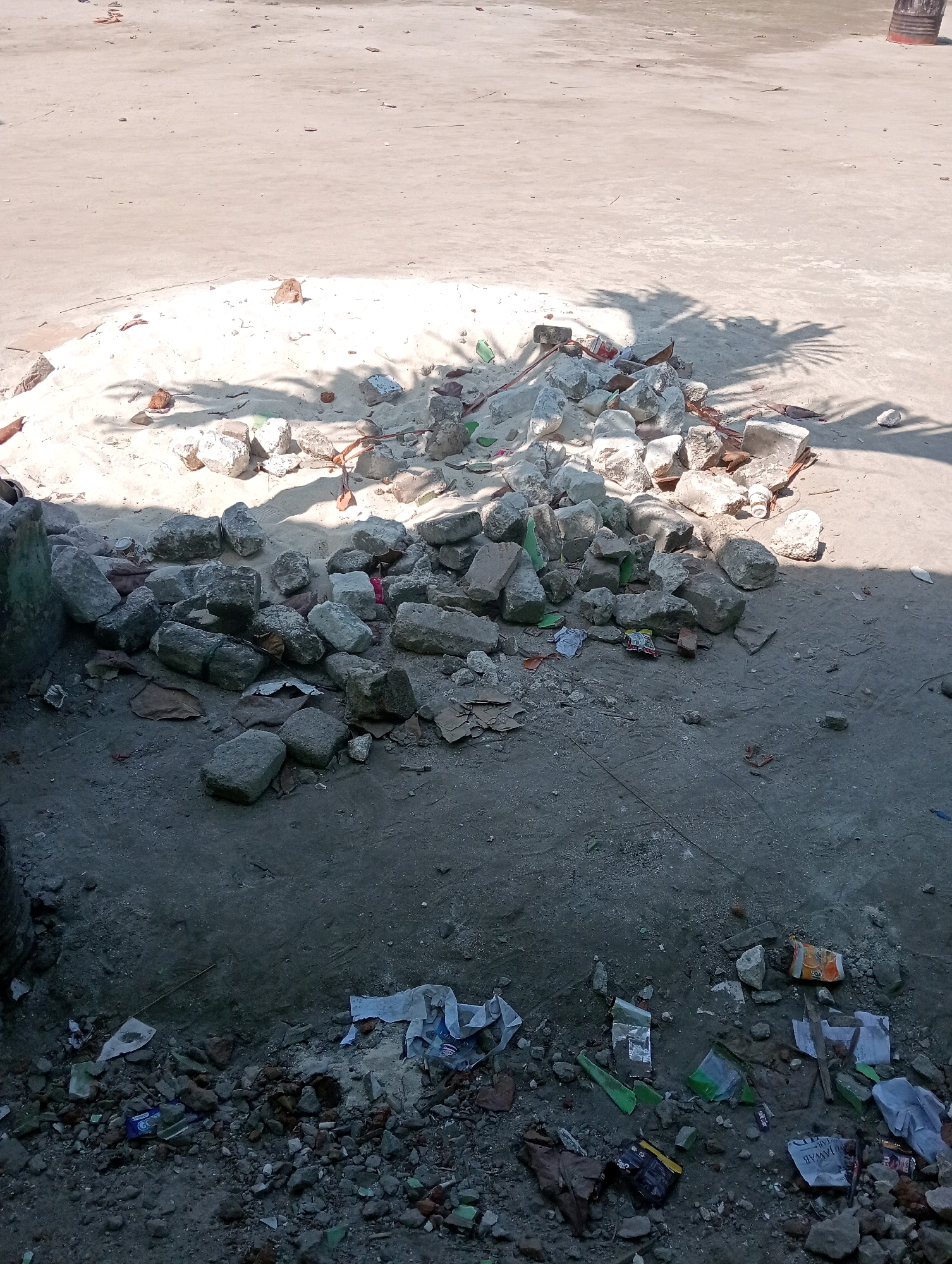 A pile of rubble and debris, consisting of concrete fragments, bricks, and scattered trash, is situated on a sun-drenched concrete surface. Shadows of palm leaves are cast across a sandy area adjacent to the rubble pile. The scene appears to be an outdoor area, possibly a construction site or a waste disposal location, given the presence of building materials and refuse. The ground is uneven, with patches of sand, dirt, and concrete. The overall impression is one of disarray and neglect.