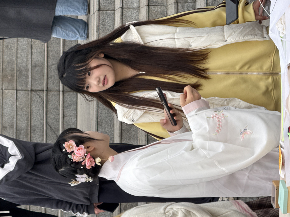 The image depicts an outdoor public scene in Jiangjin, China, focusing on two young East Asian females interacting. The individual on the left, facing right, wears a white traditional Chinese garment (Hanfu) with light pink trim on the collar and cuffs. Her dark hair is styled with multiple pink and white floral hairpins, a butterfly accessory, and a pink dangling earring. She holds a black smartphone in her left hand, its screen oriented towards the second individual. The individual on the right, facing left, has long dark hair with bangs and wears a yellow full-zip jacket under a cream puffer vest. She is engaged in conversation with the person in Hanfu. The background consists of ascending grey concrete stairs. Partially visible on the stairs are the upper torso of a third individual in a grey hooded jacket, holding a black smartphone in their left hand, and the lower torso and legs of a fourth person in a dark coat and blue jeans. In the immediate foreground, a low surface displays small boxes and white material.