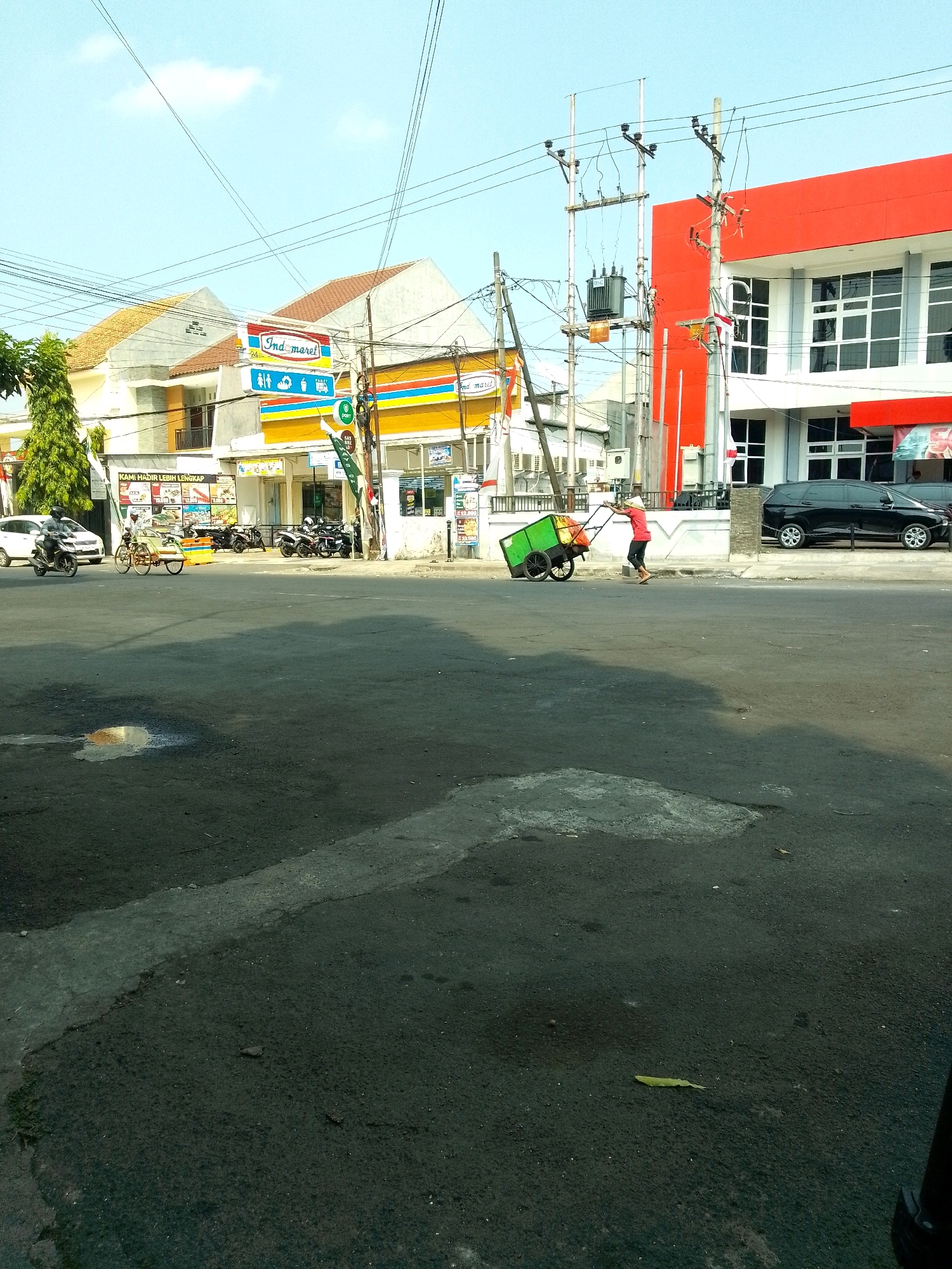 The image captures a street scene in Jember, Indonesia, under a clear sky. On the left, a white car and a motorcycle are parked. In the middle ground, a person is pushing a green cart filled with items, wearing a red shirt and a hat. Behind them, a row of shops, including an Indomaret convenience store, are visible. A bright red building stands prominently on the right side of the frame. The street is paved with asphalt, and there are puddles of water on the surface. Electrical poles and wires are present throughout the scene.
