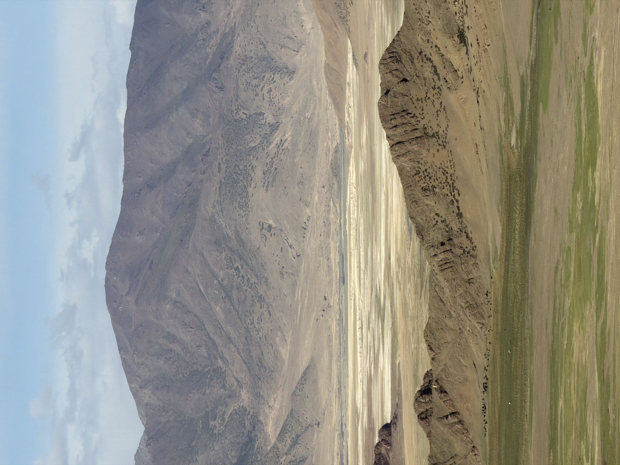 The image shows a landscape in Gyirong County, China. A large, grey mountain dominates the left side of the frame, its surface textured with ridges and sparse vegetation. Adjacent to this is a flat, sandy expanse. To the right, a rocky escarpment rises, transitioning into a field with stripes of green vegetation. The sky in the upper-left corner is pale blue with a few white clouds. The image is rotated.