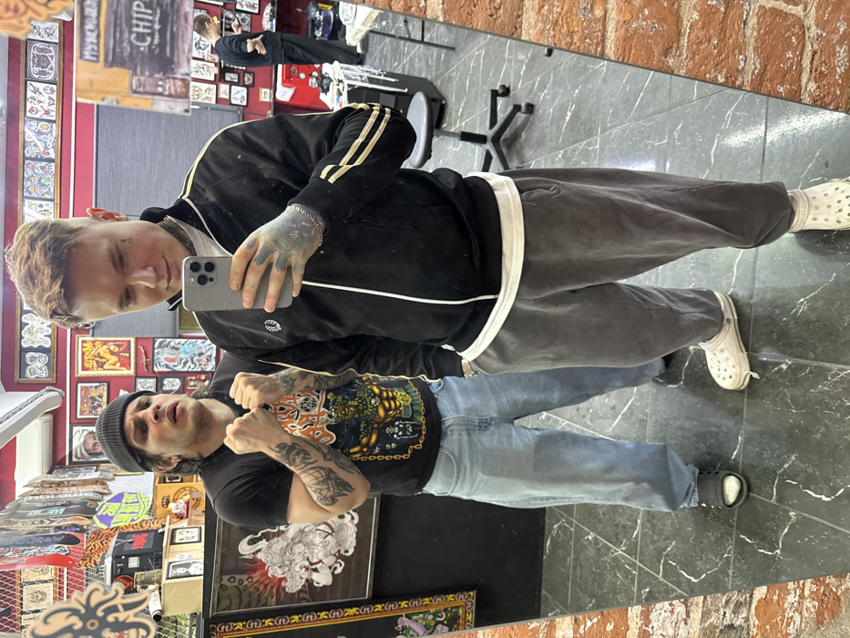 Two men are reflected in a mirror. The man in the top position takes a selfie with an iPhone. He wears a black jacket, gray sweatpants, and white Crocs. Tattoos adorn his neck and hands. Below, the second man, wearing a black beanie, black T-shirt, and blue jeans, clenches his fists. Tattoos also cover his arms. Behind them, the room has a red wall filled with tattoo art, skateboards, and various decorations. A glimpse of another person can be seen in the mirror, standing by a tattoo chair. The floor is gray marble.