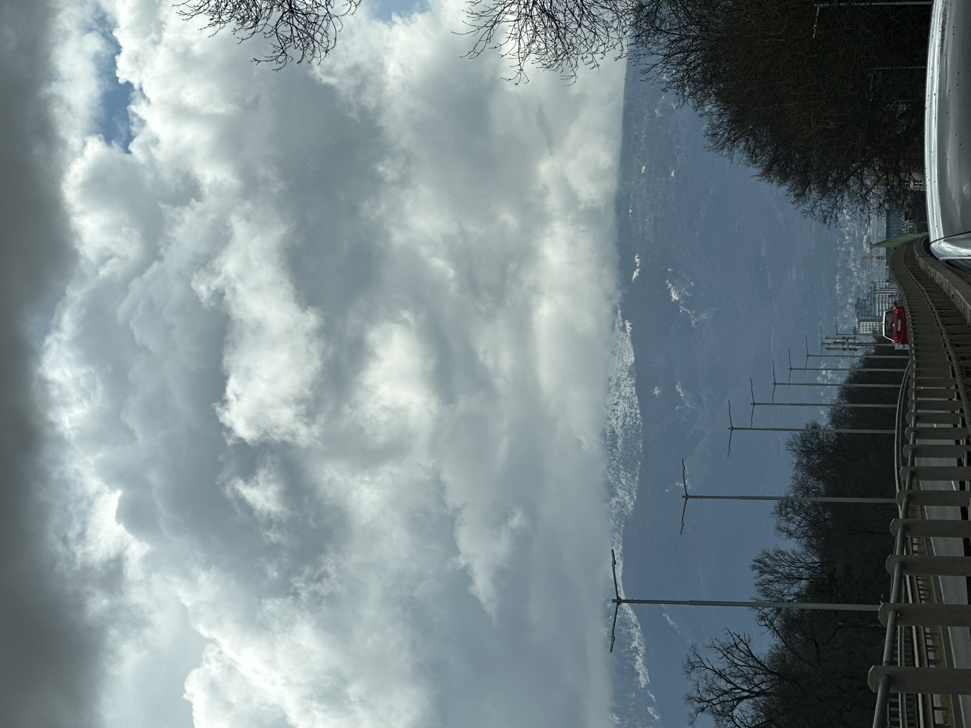 The image depicts a view from a road or bridge, possibly in Sofia, Bulgaria. In the foreground, a metal guardrail runs alongside the road. Beyond the guardrail, there are streetlights and trees, some bare, indicating a possibly cooler season. A red car is visible further down the road. In the background, there's a mountain range, some parts of which appear to be covered with snow or ice. The sky is filled with predominantly gray clouds, suggesting an overcast day. A glimpse of blue sky can be seen through a break in the clouds. The photo appears to have been taken from inside a car, as a portion of the car is visible on the right side of the image. The scene depicts an urban landscape merging with a natural mountain setting. Given the presence of the mountain range, the location is likely in or around Sofia, Bulgaria.