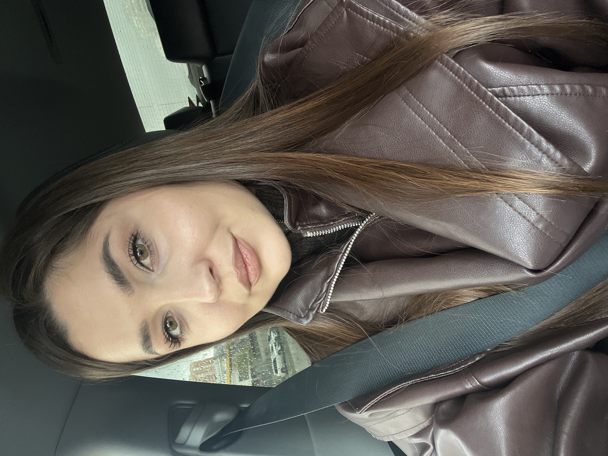 A young woman is in the driver's seat of a car. She has long brown hair and hazel eyes. She is wearing a brown leather jacket and a black turtleneck sweater. The car seatbelt is fastened. Raindrops are visible on the car window, suggesting it is raining outside. The location is likely not Moscow, Russia, as there are no elements in the image to indicate that.