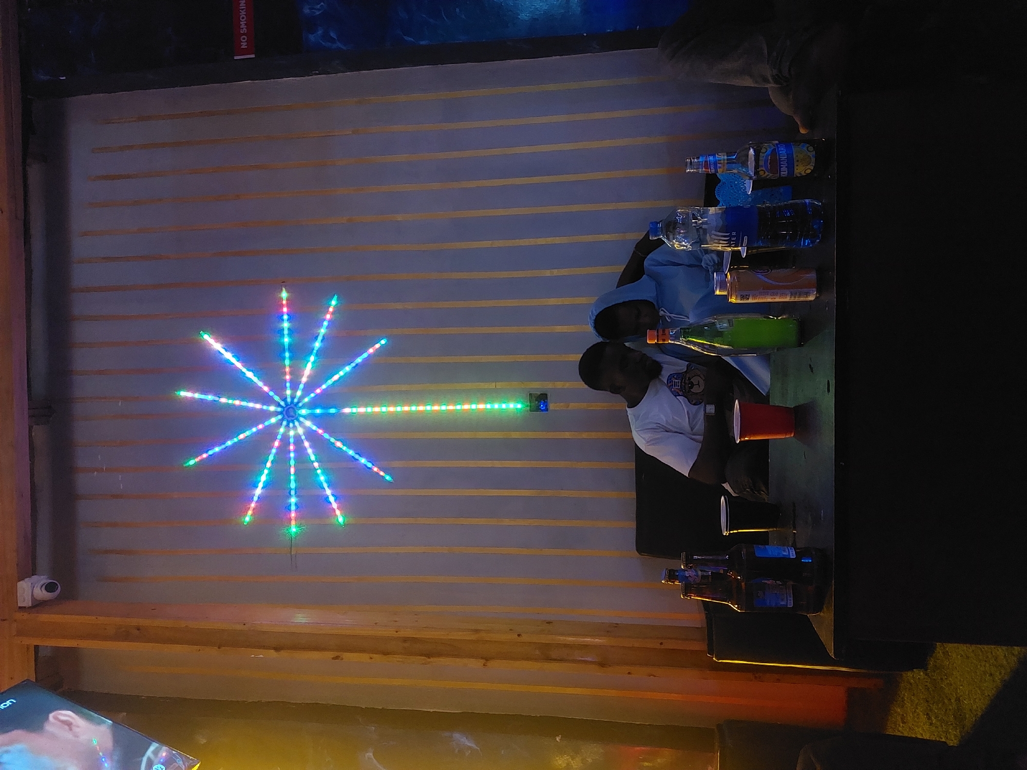 The image shows an indoor setting, possibly a bar or lounge, featuring two individuals and several bottles of beverages. The background wall is decorated with horizontal yellow stripes and a starburst-shaped light fixture with multiple colored LED lights. A surveillance camera is mounted on the left side of the wall.
Two individuals are visible; one is wearing a light blue hooded sweatshirt and the other is wearing a white t-shirt. A red plastic cup sits on the table in front of the person in the white shirt. There are multiple bottles on the table, including a water bottle with a blue cap and other bottles containing liquids of different colors.
Based on the bottles present, some of the beverages may include water, juice, and alcohol. A black cup and bottles sit on the table.
The location in Dar es-Salaam, Tanzania, is unknown from the image.