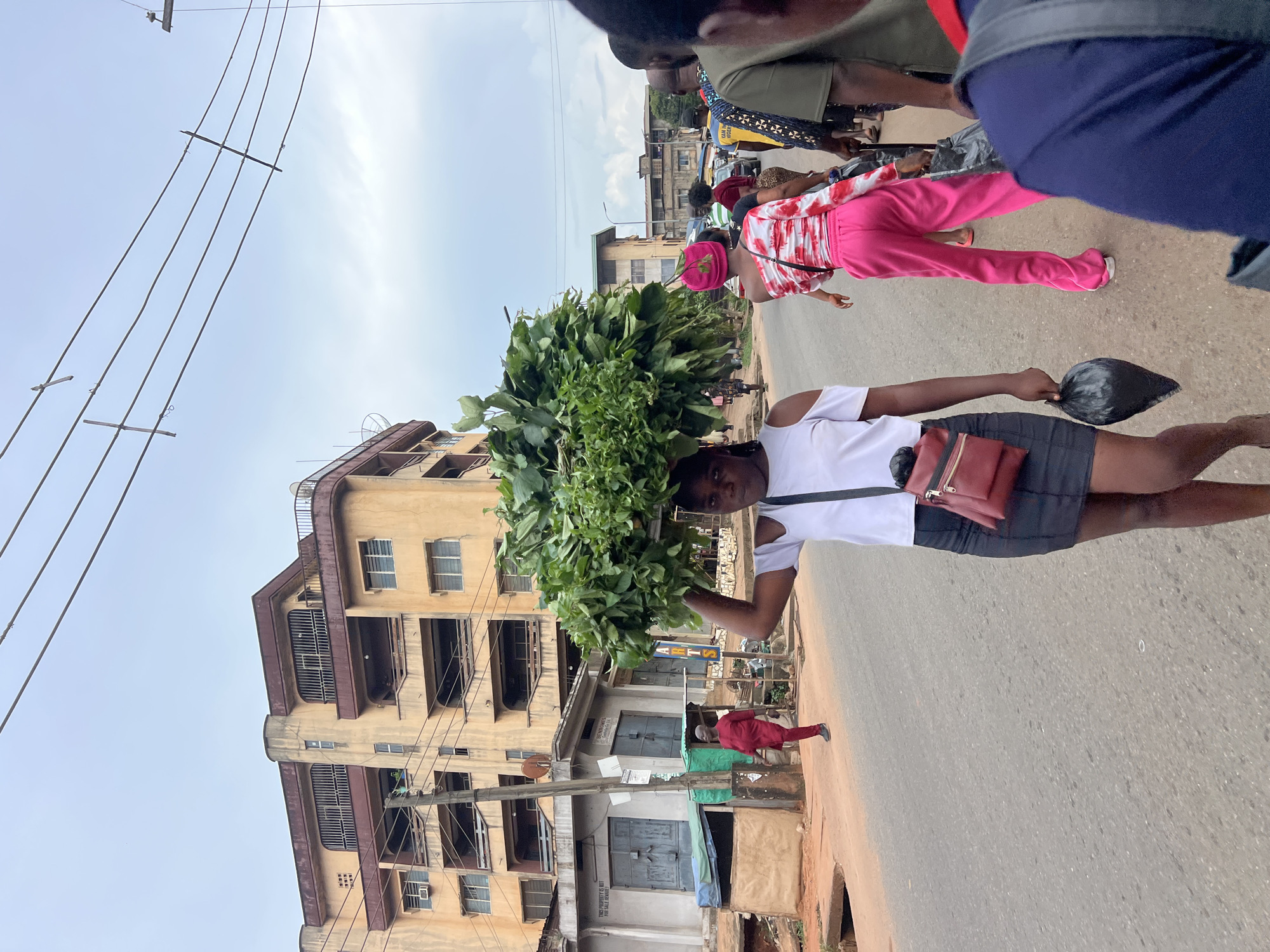 The image shows a street scene in Nkpor, Nigeria. In the foreground, a young woman carries a large bundle of green leafy vegetables on her head. She is wearing a white off-the-shoulder top, a black skirt, and a red crossbody bag. To her left, a man in a red traditional outfit is walking away from the camera. Behind her, another woman is walking in the same direction, wearing a pink and white patterned top and bright pink pants, with a pink headscarf. Several other people and vehicles are visible further down the street. The background features a multi-story building with balconies and windows, and a cloudy sky overhead. Power lines are strung across the sky. The environment appears to be a busy urban area with a mix of commercial and residential buildings.