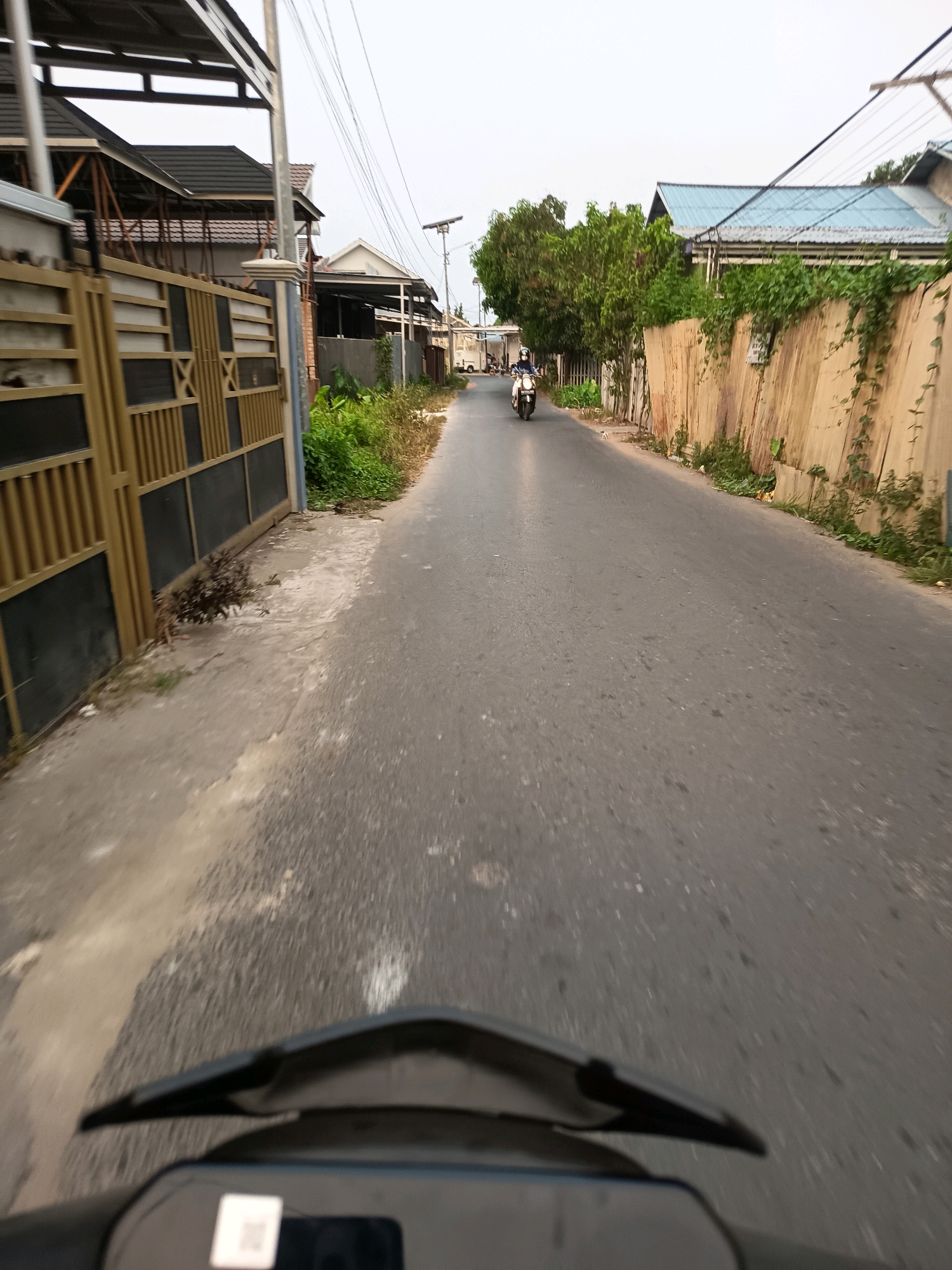 The image captures a street scene in Banjarmasin, Indonesia, likely during daytime. The perspective is from a moving vehicle, possibly a motorcycle, as the handlebars and part of the dashboard are visible in the foreground. The road is asphalt and appears to be in moderate condition with some visible patches and cracks.
On the left side of the road, there is a property with a golden and black metal gate and fence, followed by houses with visible roofs and structures. Green vegetation and some plants are present along the fence line. On the right side of the road, there is a tall wooden fence, possibly made of planks, with greenery and vines growing on it. Further down the road, more houses with distinct blue roofs can be seen, along with trees and utility poles carrying electrical wires.
A person on a white motorcycle is visible in the distance, traveling on the road. The sky is overcast, suggesting cloudy weather. The overall impression is of a residential street in an urban or suburban area of Banjarmasin.