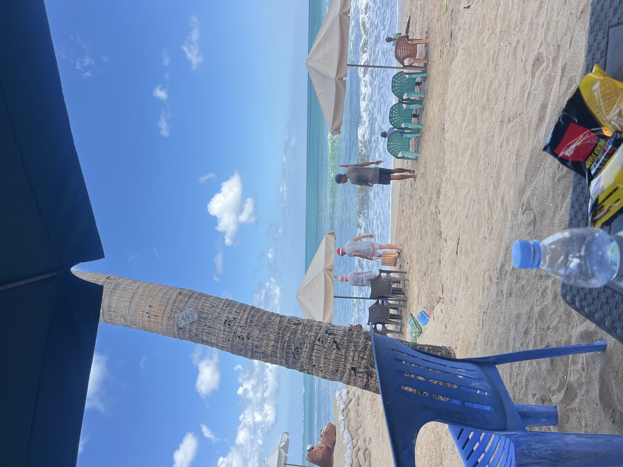 This is a photograph taken at Kuta Beach, Bali, Indonesia. The image captures a sunny day with a clear blue sky dotted with white clouds. In the foreground, a blue plastic chair is positioned on the sandy beach, with a table in front of it holding a plastic water bottle and a bag of snacks.
Beyond the chair, the beach extends to the ocean. A large palm tree trunk stands prominently to the left of the frame, partially covered by a dark umbrella. Several beach umbrellas are set up on the sand, providing shade.
People are visible in the middle ground. Two individuals, one wearing a red hat and the other a light-colored headscarf, are standing near a beige umbrella. Further down the beach, a man in a gray t-shirt and shorts is walking away from the camera. In the distance, several green plastic chairs are arranged under an umbrella, with some people sitting on them. The ocean waves are rolling onto the shore.
In the background, more beach umbrellas and sandbags are visible, as well as a person sitting on a beanbag chair under an umbrella. The overall scene depicts a relaxed atmosphere at a popular tourist beach.