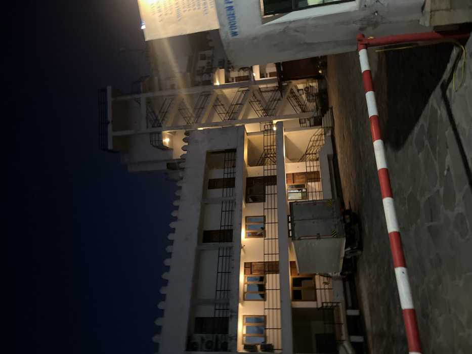 The image depicts a multi-story building at night, illuminated by artificial lights against a dark, unlit sky. The building, likely made of light-colored (possibly white or off-white) material, features distinct architectural elements.
The main building on the left has several floors with rectangular windows, some of which are covered by dark metal grates or bars. Many windows are lit from within, casting a warm yellow-orange glow. The facade also has prominent vertical projections, giving it a somewhat serrated or stepped appearance on its left edge. Balconies or walkways extend from the upper floors, featuring decorative metal railings that form a repeating V-shaped pattern. External staircases with metal railings are also visible, suggesting access between floors from the outside. Air conditioning units are mounted on the lower part of the building.
In the mid-ground, next to the building, a piece of lifting equipment, resembling a cherry picker or a boom lift, is parked. It has a gray, caged platform with yellow and black hazard stripes, suggesting recent or upcoming maintenance or construction work.
On the right side of the foreground, a red and white striped boom barrier or gate pole stands upright, indicating an entrance or restricted access area. The ground beneath it and extending into the mid-ground is paved with irregularly shaped, light-colored stones or tiles.
A bright light source, likely a streetlamp, is visible in the upper left portion of the image, creating a strong lens flare and illuminating parts of the dark sky.
A large, white banner or sign hangs from a higher part of the building, partially obscured by the bright light. Visible text on the sign, in dark lettering, includes:
- On the left side: "SIT", "LADIES", "CHILDREN", "CHINESE", "CHOPSTICK", "CHICKEN", "FRIED", "SPECIAL", "CHILLI".
- On the right side: "WOODEN P A L..." (likely part of "WOODEN PALACE" or similar).
The overall scene is captured at night, with the artificial lighting creating strong contrasts and highlighting the architectural details and the operational nature of the location, possibly a hotel, restaurant, or residential complex.