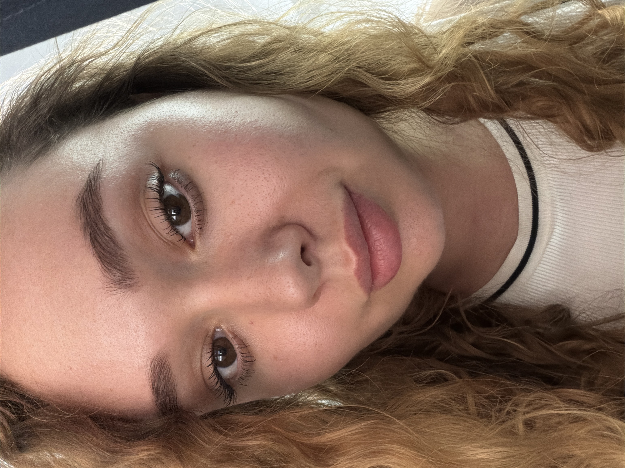 The image shows a close-up of a young woman's face. She has fair skin, brown eyes, and naturally curly, blonde hair. She is wearing a white sweater with a black stripe. Her eyebrows are well-defined and slightly arched. She has full lips. The background appears to be a light-colored surface. There is no information available in the image to place this image in the city of Ramat Gan, Israel.